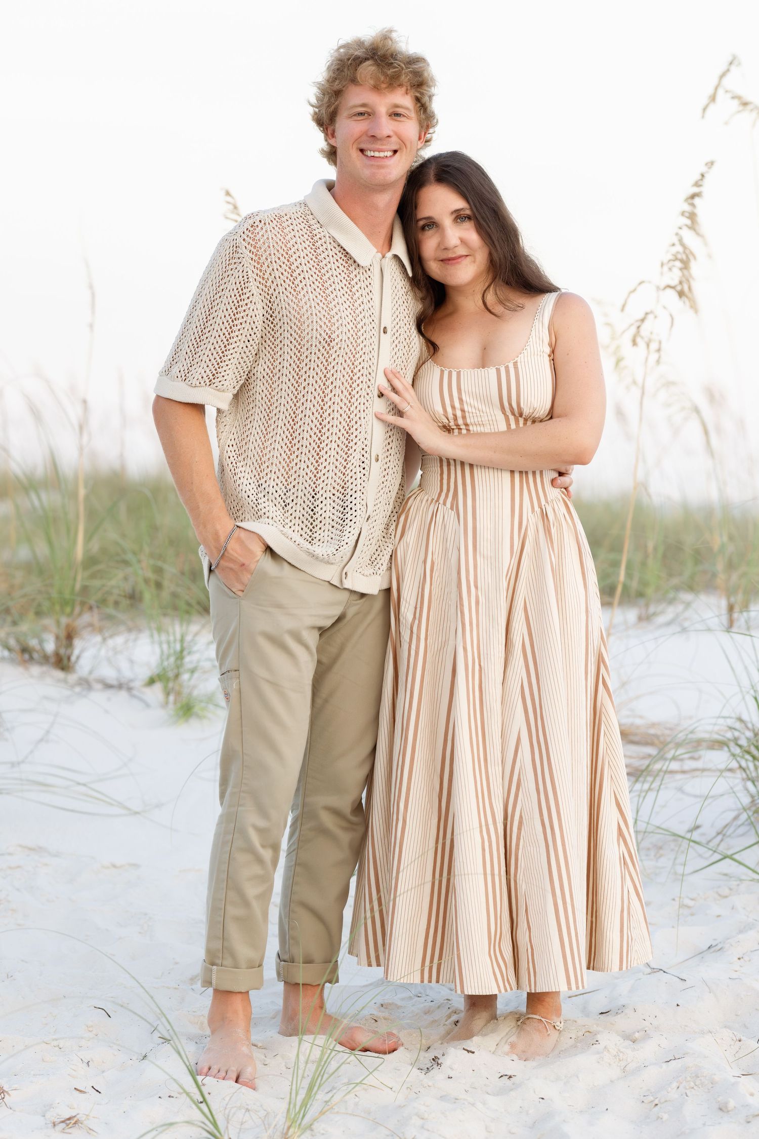 Couple embracing on sandy beach, man in crochet shirt and khakis, woman in striped maxi dress, sea oats background.