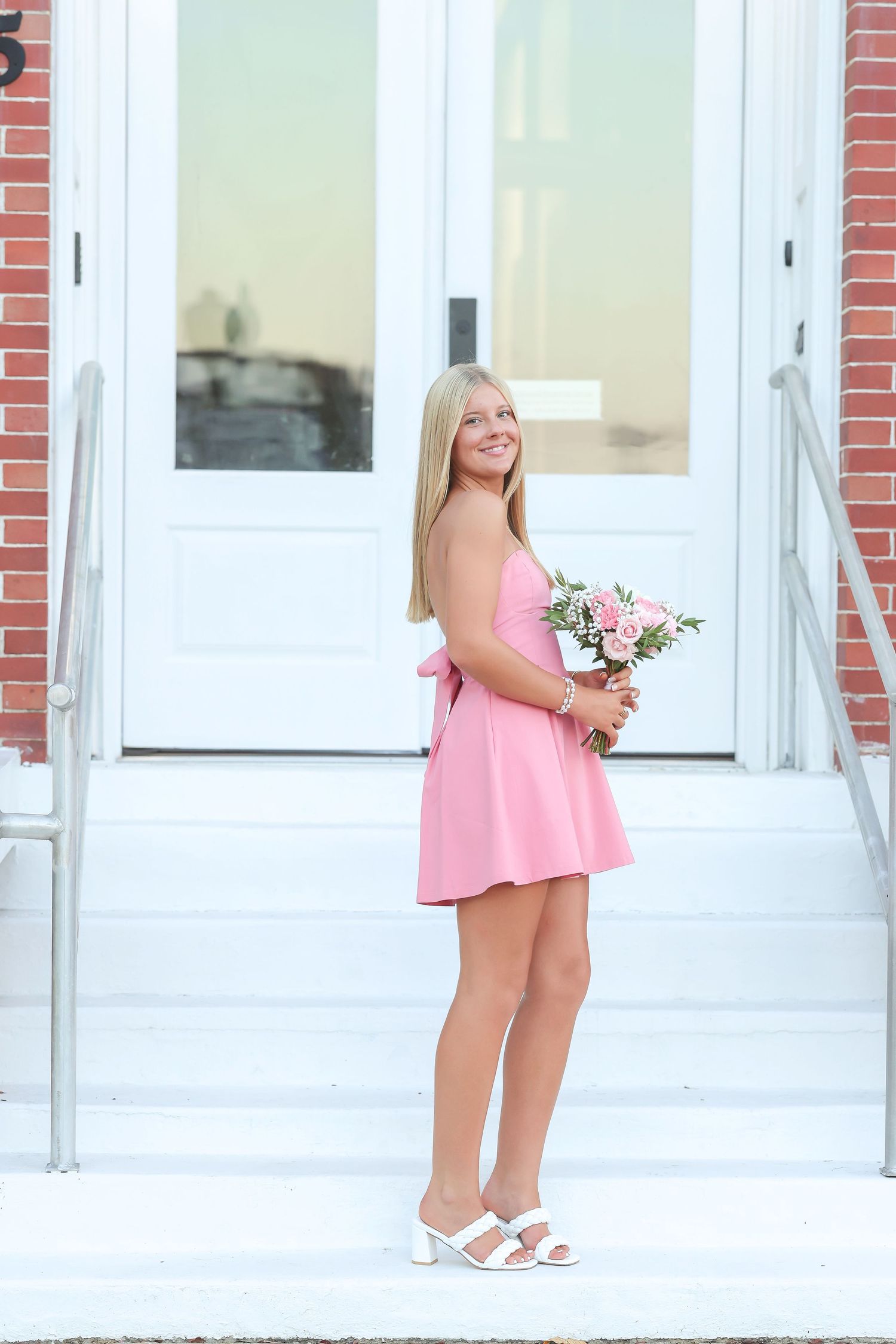 A person in a pink dress holds a floral bouquet against a white columned building exterior.