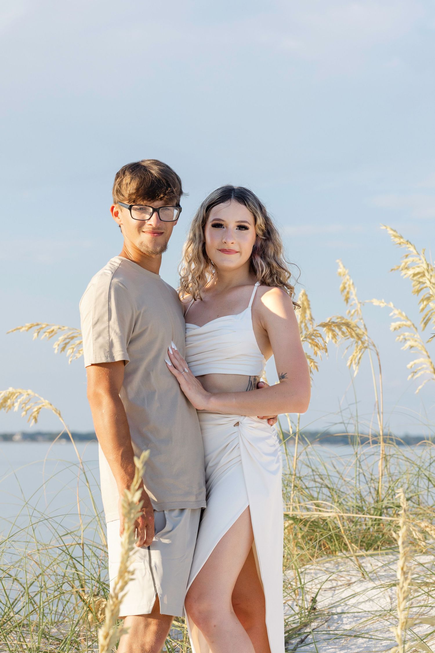 A couple poses together on a beach with sea oats in the foreground on a sunny day.