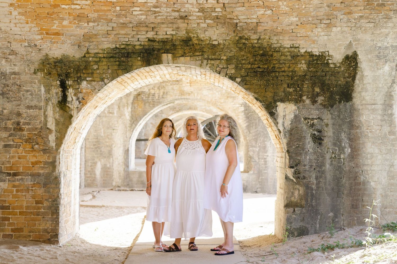 Three people in white dresses standing under stone archway at historic fort.