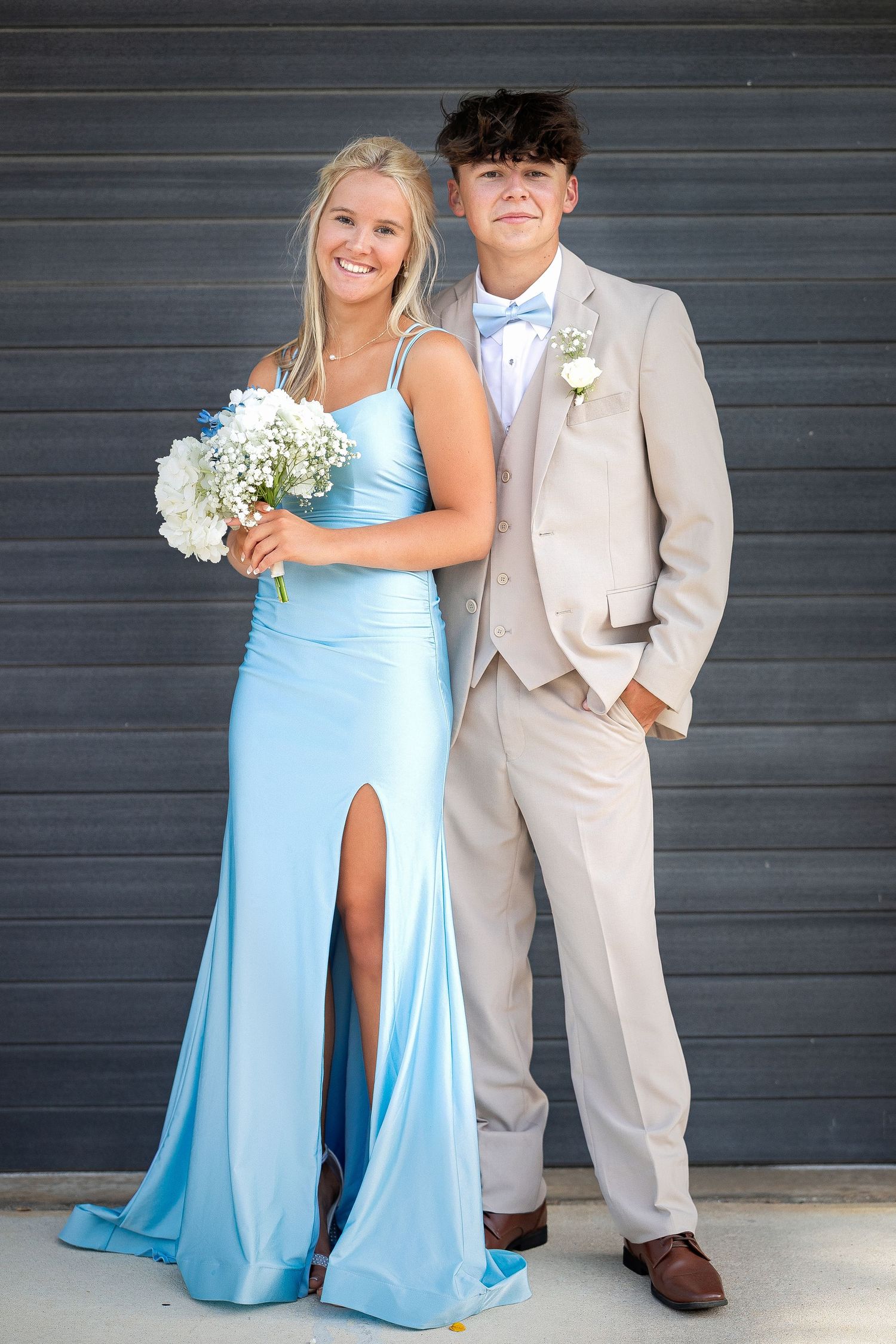 Couple in formal attire pose for prom photo with light blue dress and beige suit against gray wall background.