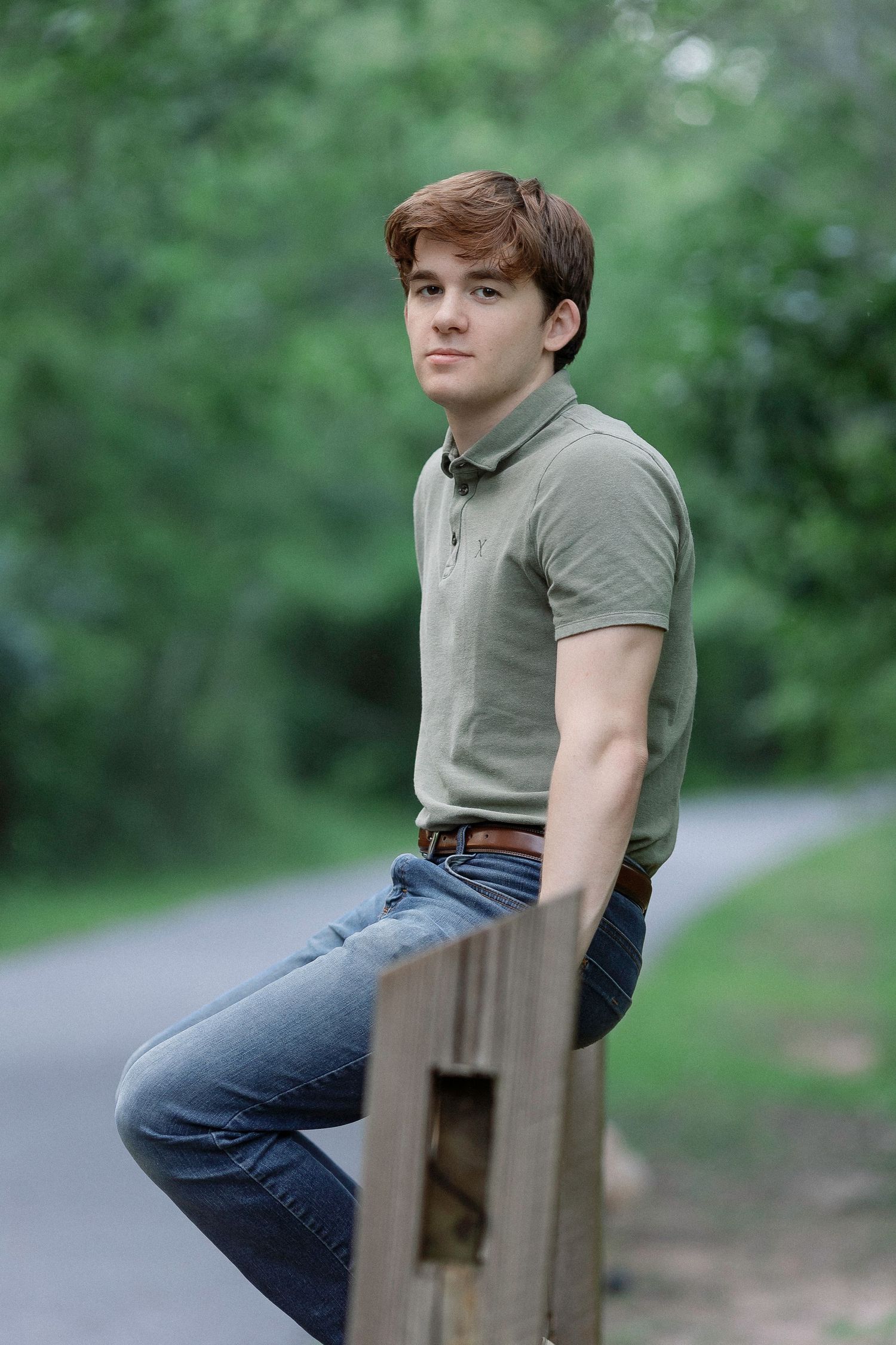Young man in green polo shirt and jeans sits on wooden fence along tree-lined path outdoors.