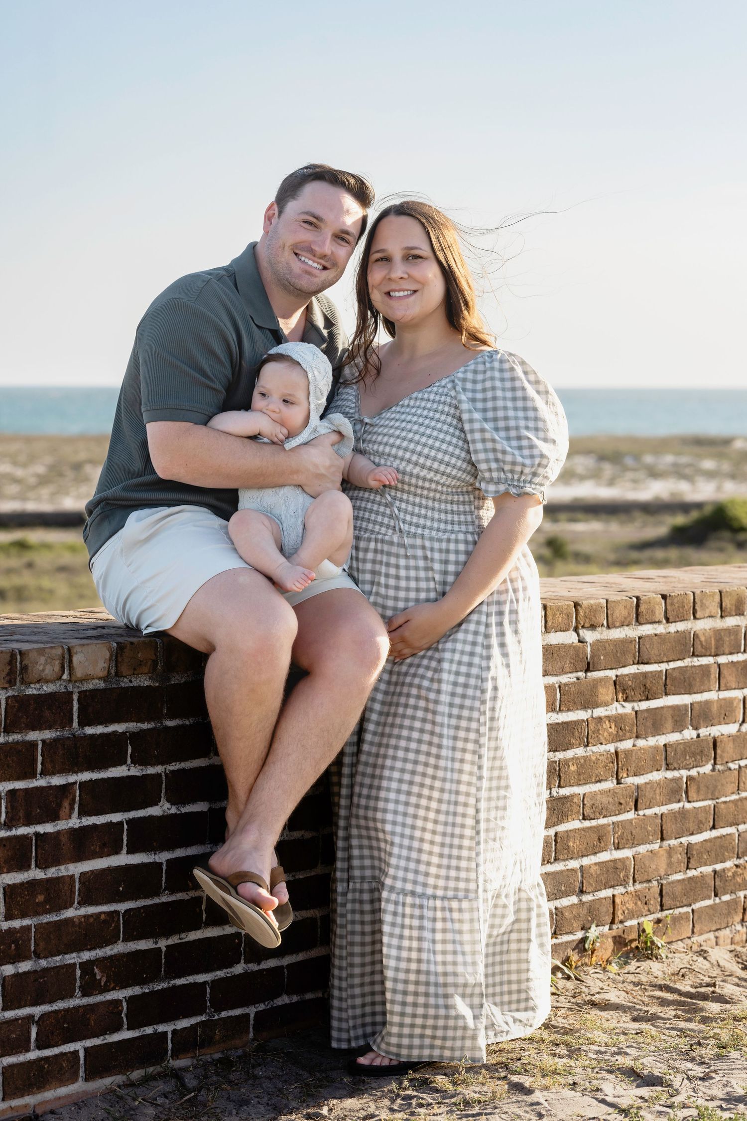 A smiling couple with their baby sits on a brick wall at the beach on a sunny day.