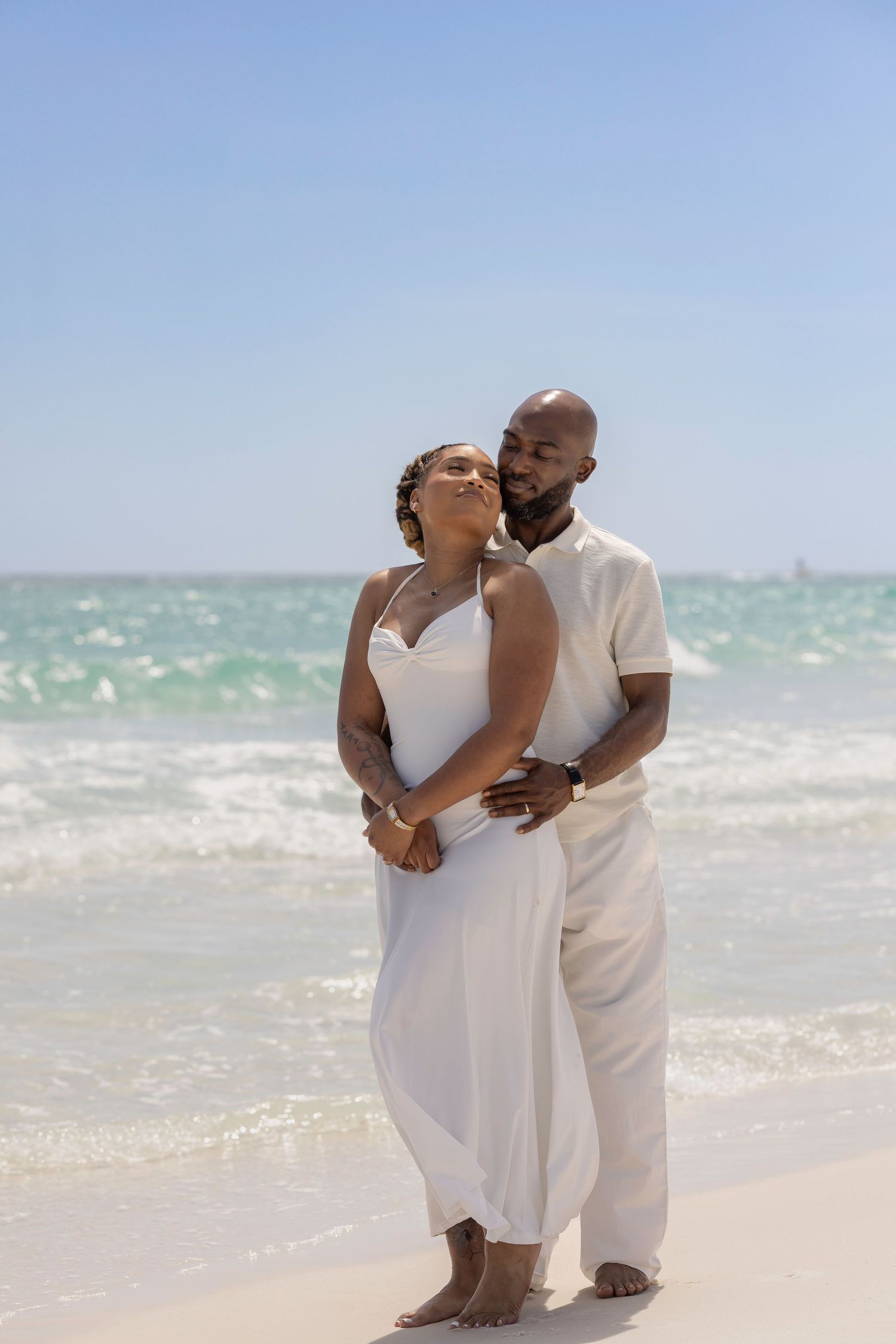 A couple in white outfits embraces on a sunny beach with turquoise waves behind them.