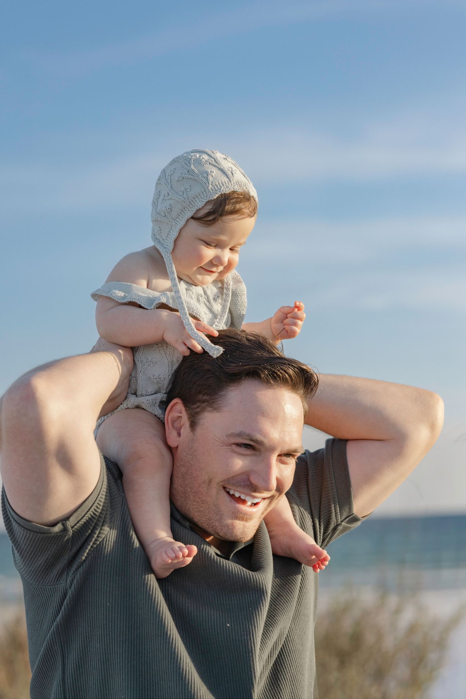 Happy father carrying smiling baby on shoulders at the beach on a sunny day.