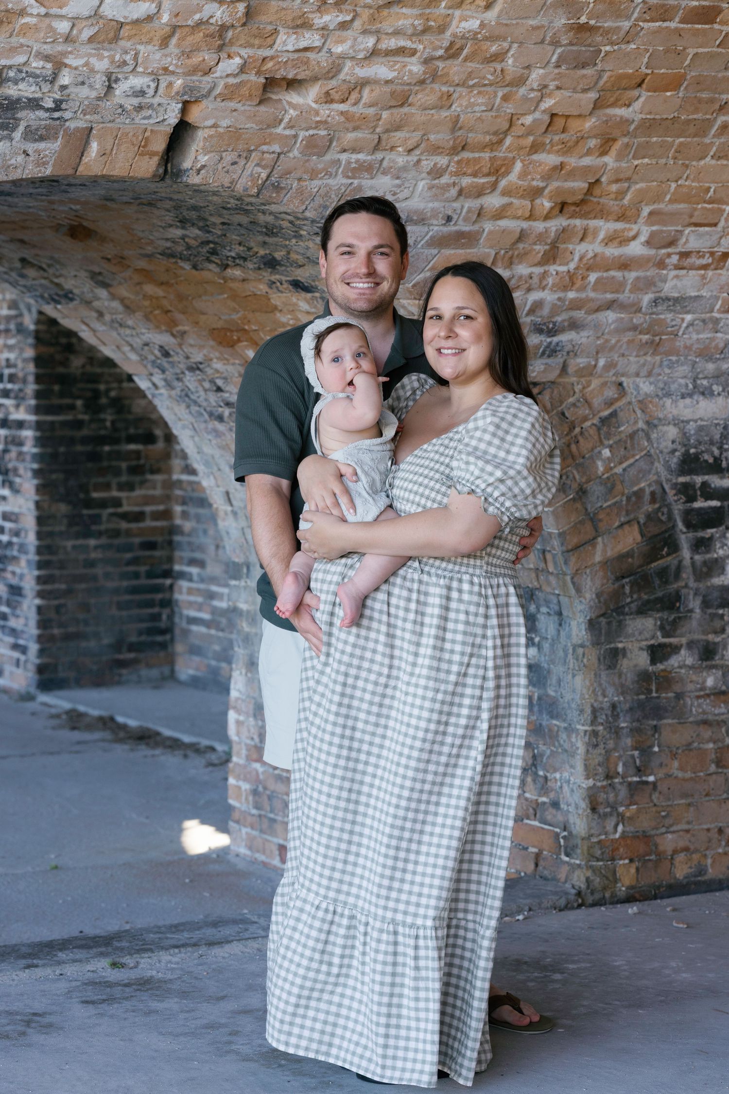 A happy family of three poses under a brick arch, with parents holding their baby girl in a checkered dress.