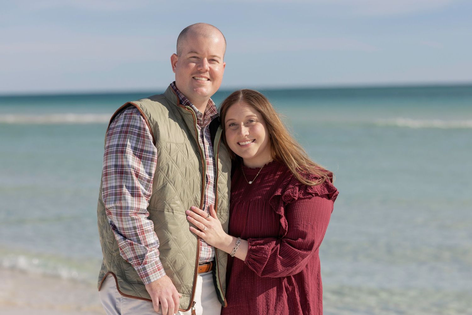Happy couple poses together on a sunny beach with turquoise ocean water in the background.