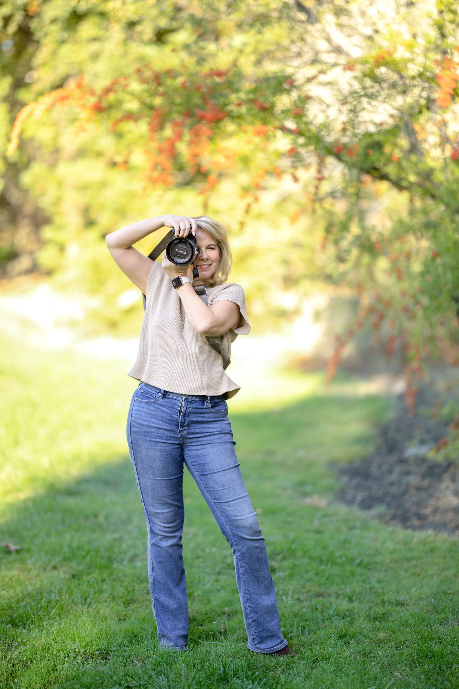 Photographer with camera stands on grassy lawn capturing photos against blurred autumn foliage background.