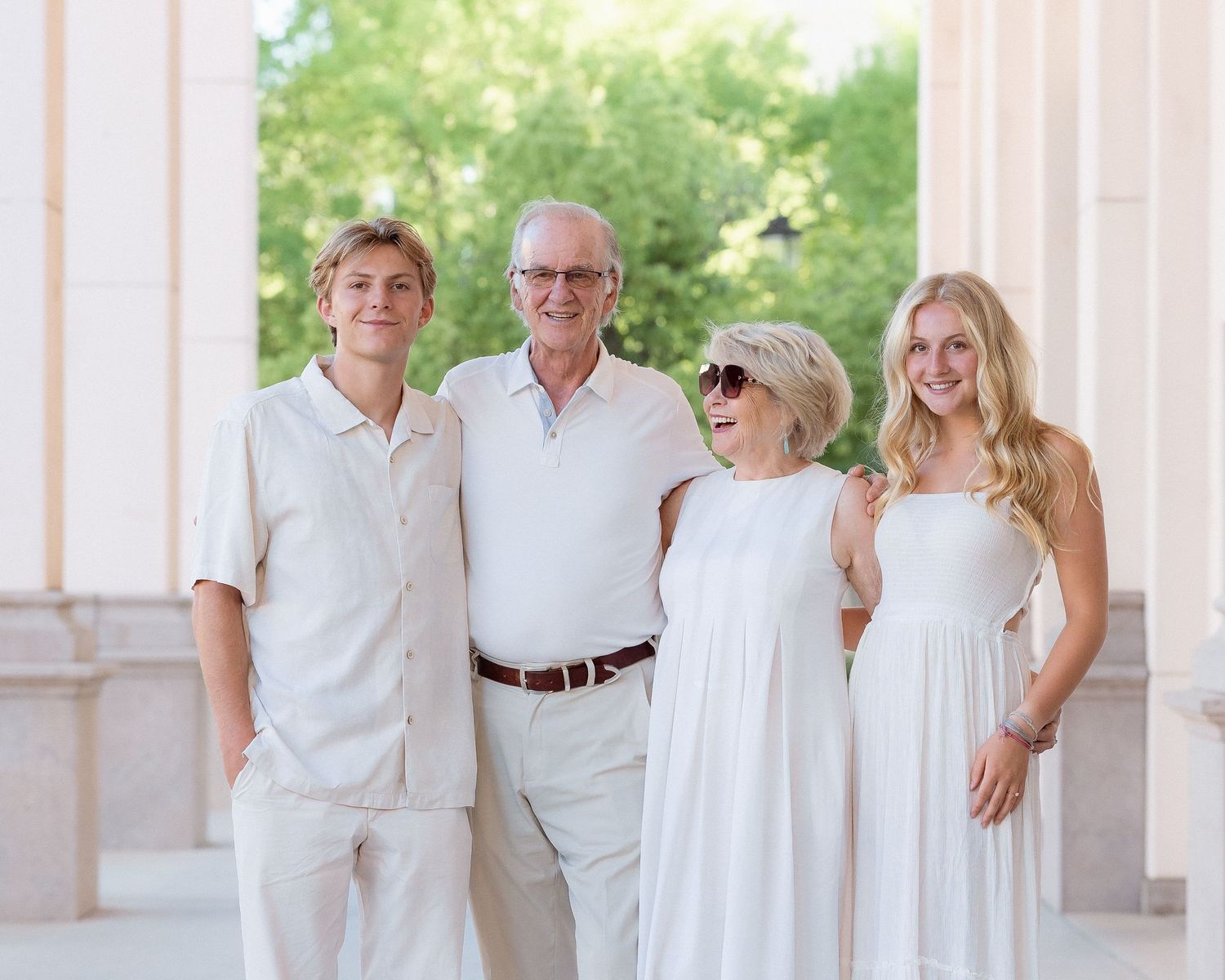 Family of four posing in white outfits outdoors near elegant columns on a sunny day.