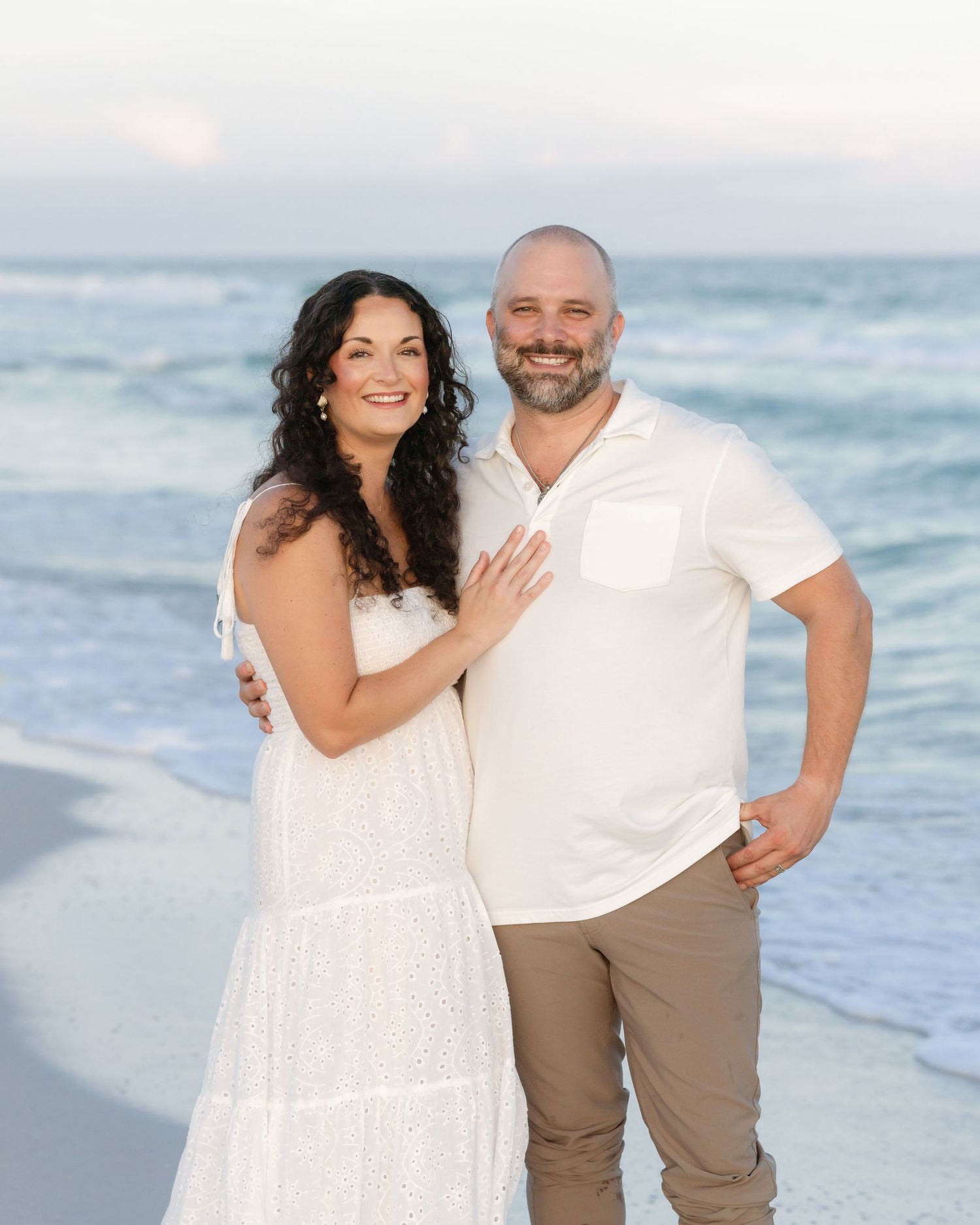 Couple in white outfits smiling together on a sunny beach with ocean waves behind them.
