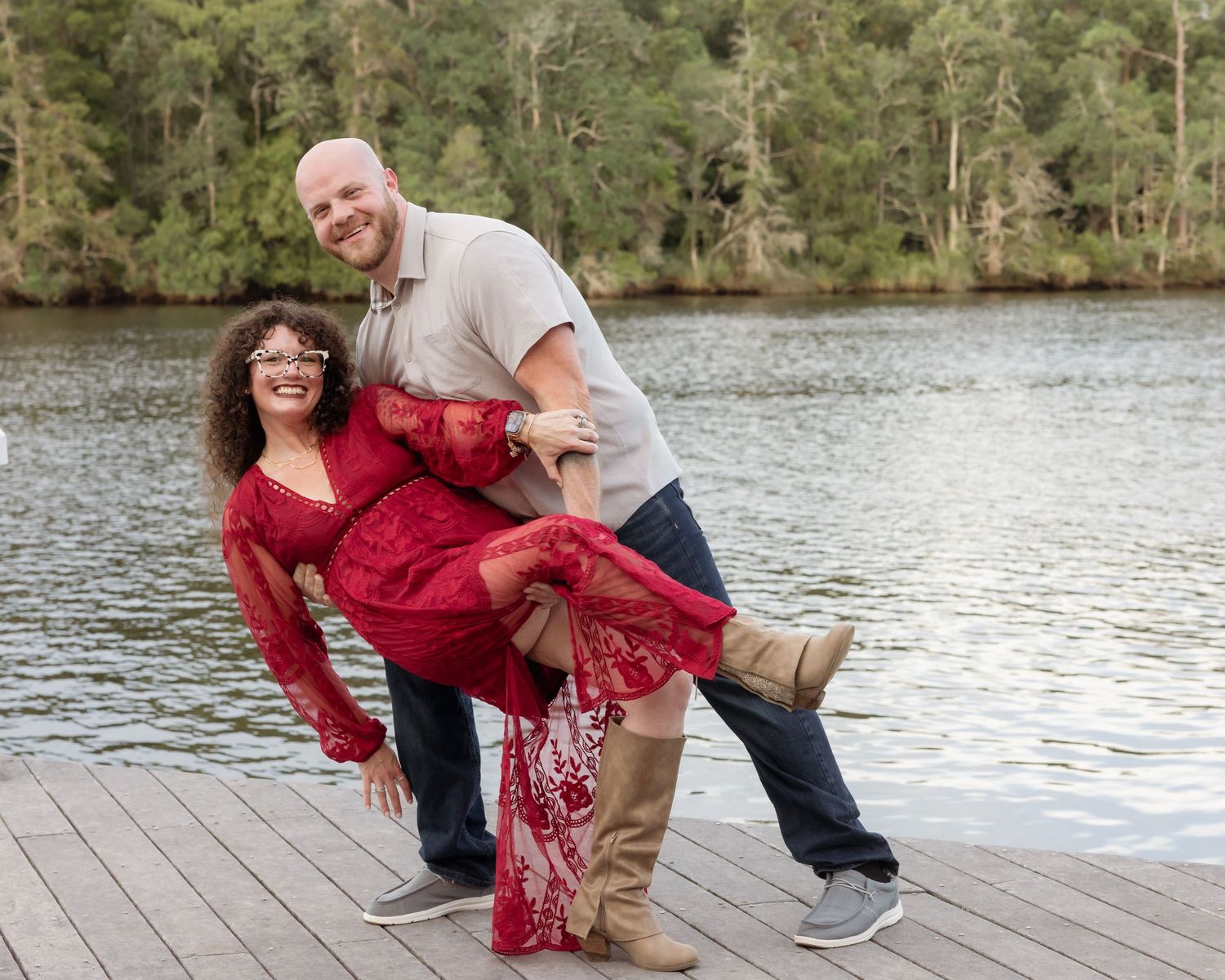 Joyful couple posing on a lakeside dock, woman in red lace dress dipping playfully with her partner in casual attire.