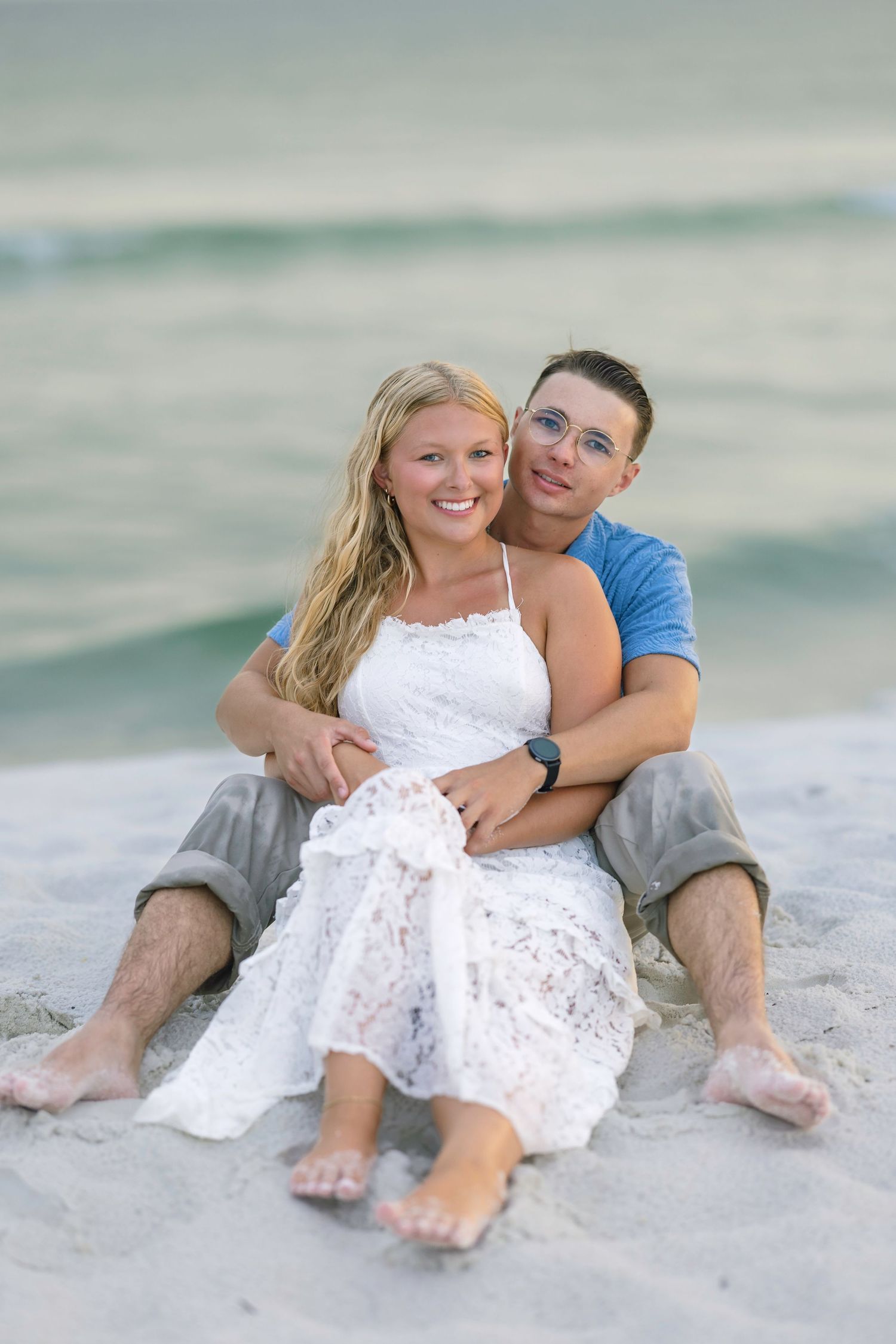 A happy couple sits together on a sandy beach, woman in white lace dress leaning back into mans arms near the ocean.