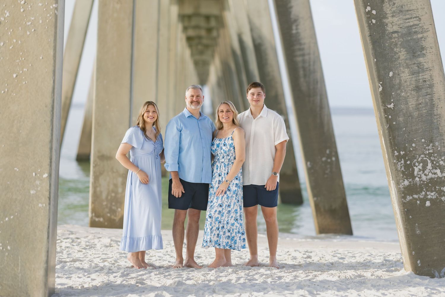 Family of four smiling together under a beach pier on white sand with ocean views in the background.
