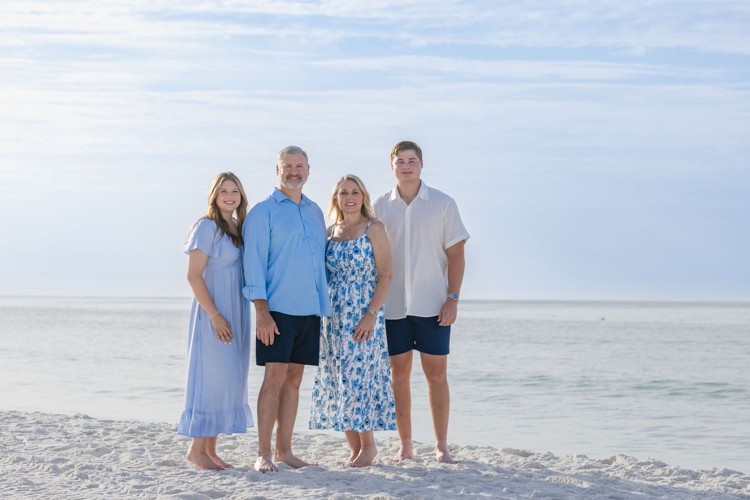 Family of four posing together on a sandy beach with calm ocean waters and blue sky in the background.
