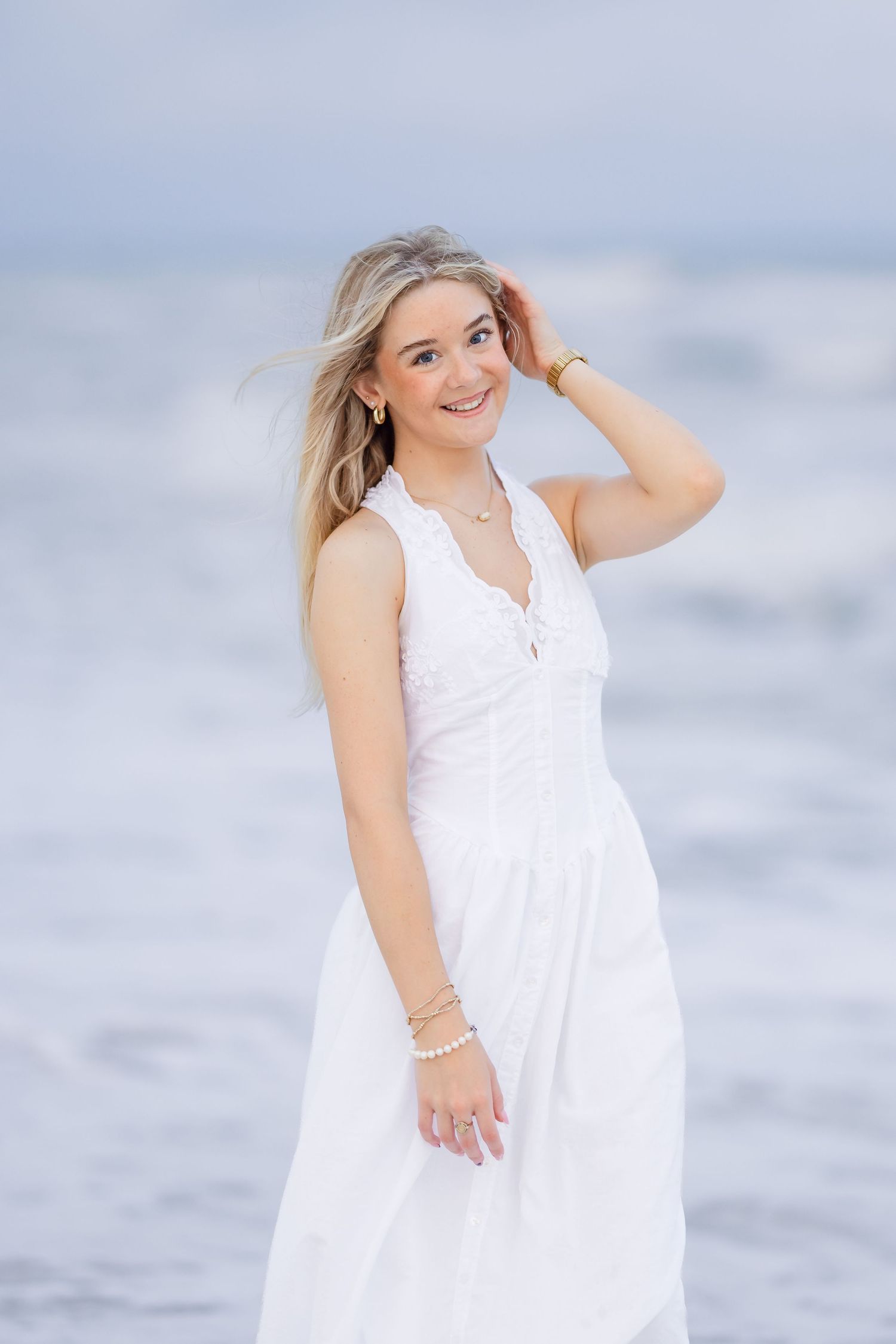 Young blonde woman in white sundress smiling at beach, wearing gold jewelry, with ocean waves in background.