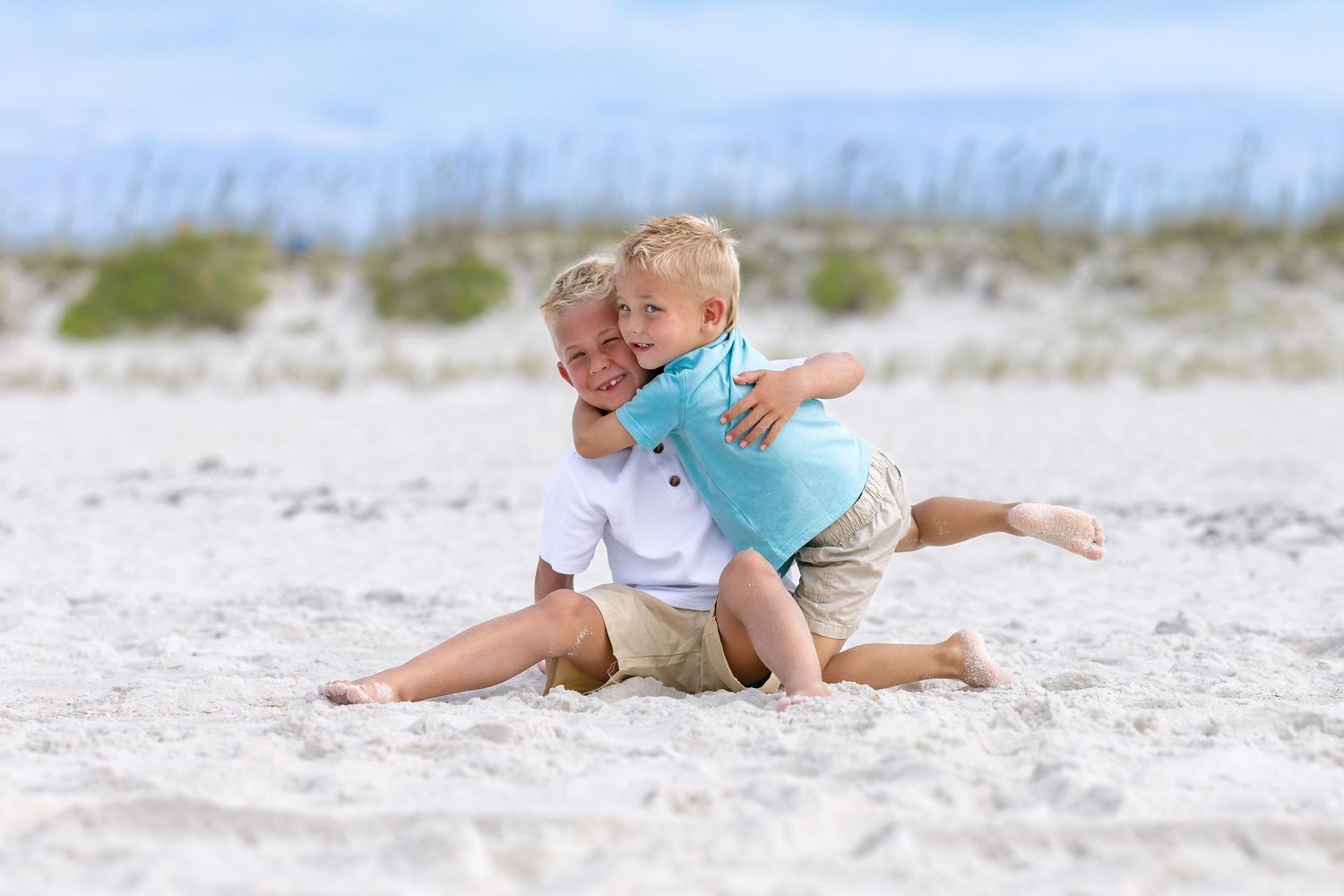 Two young boys playing and hugging on a white sandy beach on a sunny day.