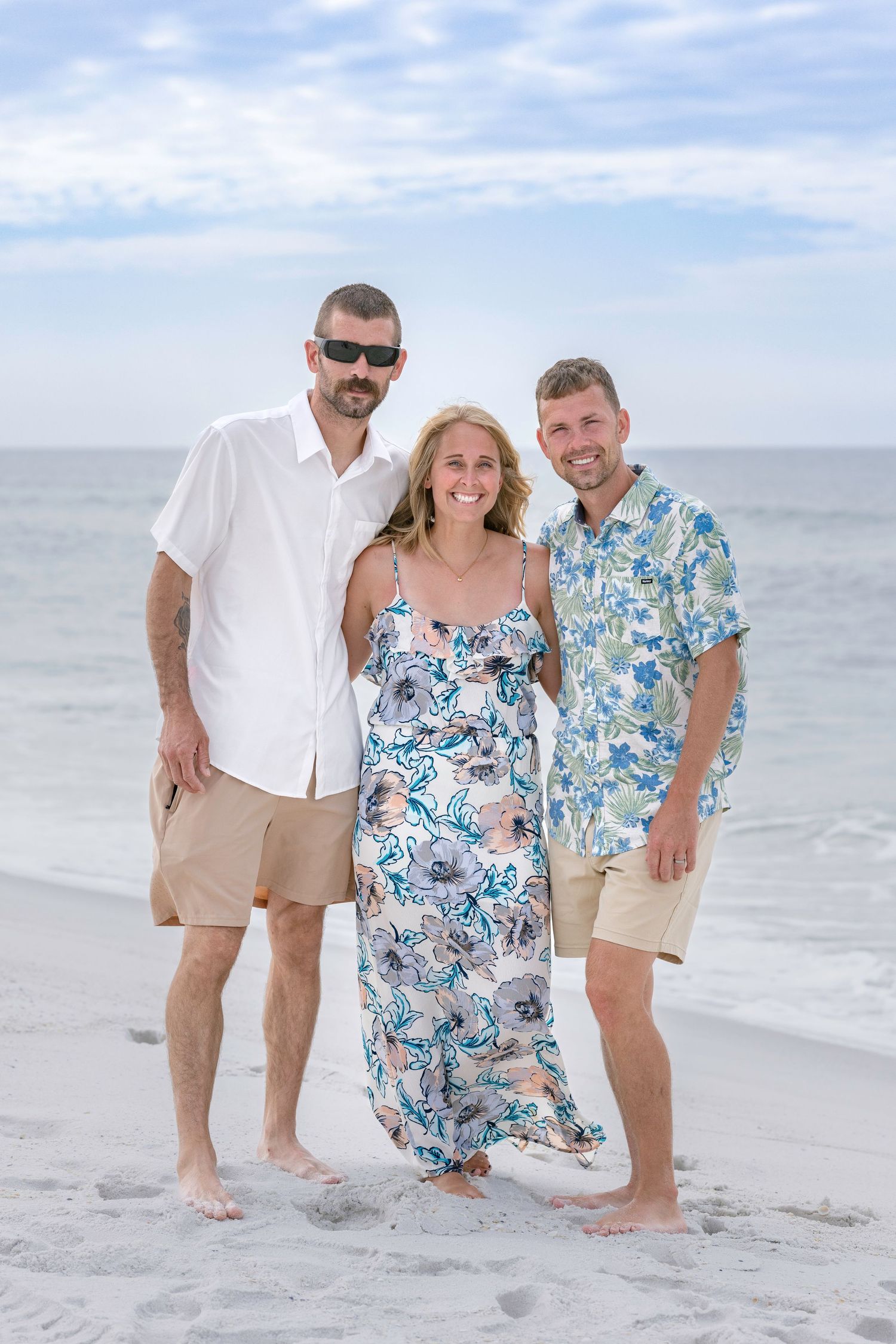Three smiling adults pose together on a sandy beach with ocean waves in the background on a cloudy day.