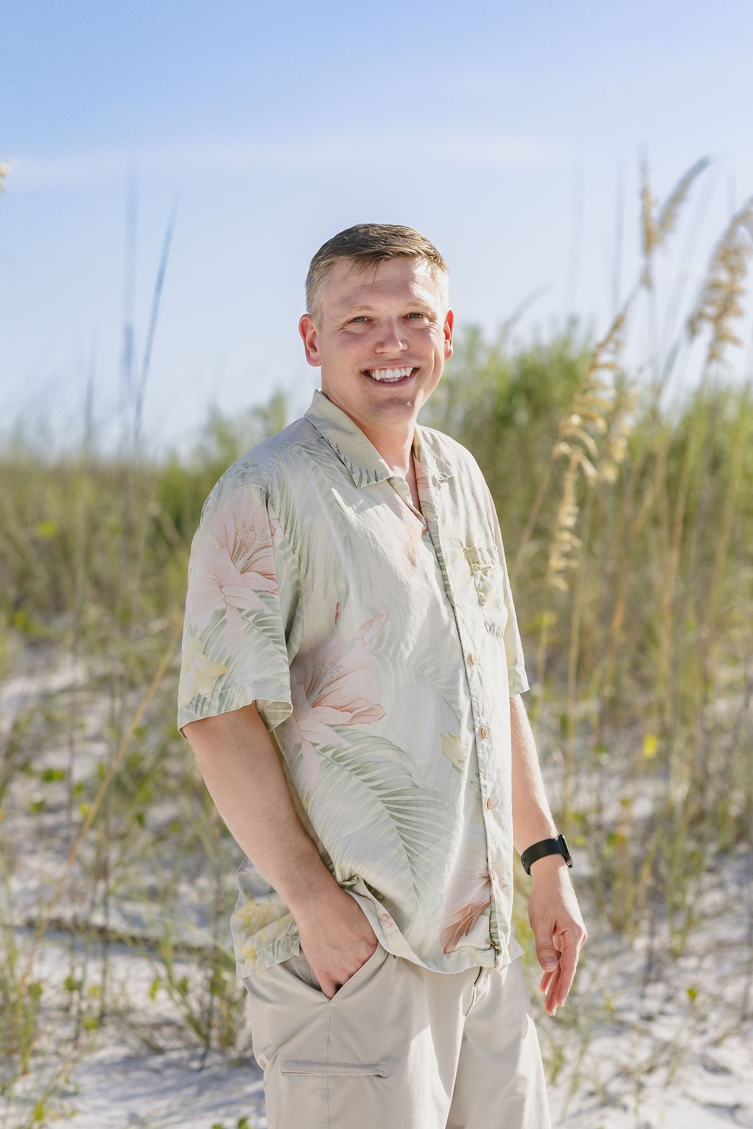 Smiling man in floral shirt standing on sandy beach dunes with sea oats and blue sky background.