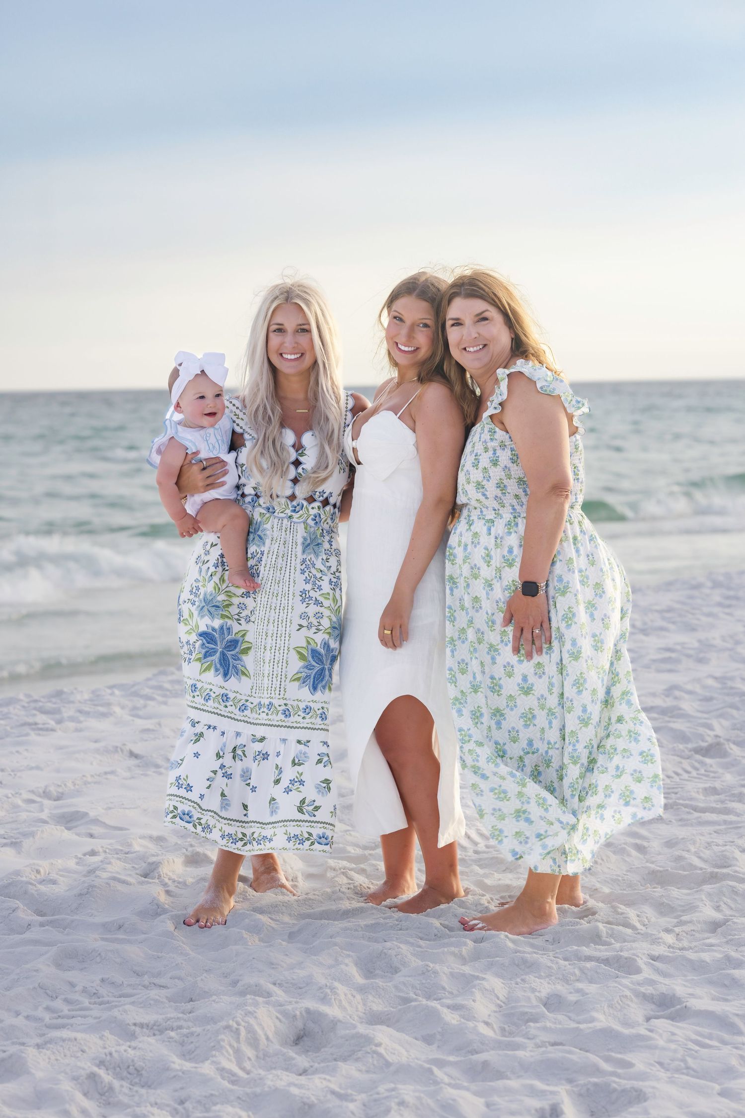 Three women in floral dresses pose together on a sandy beach, one holding a baby girl with a white bow.