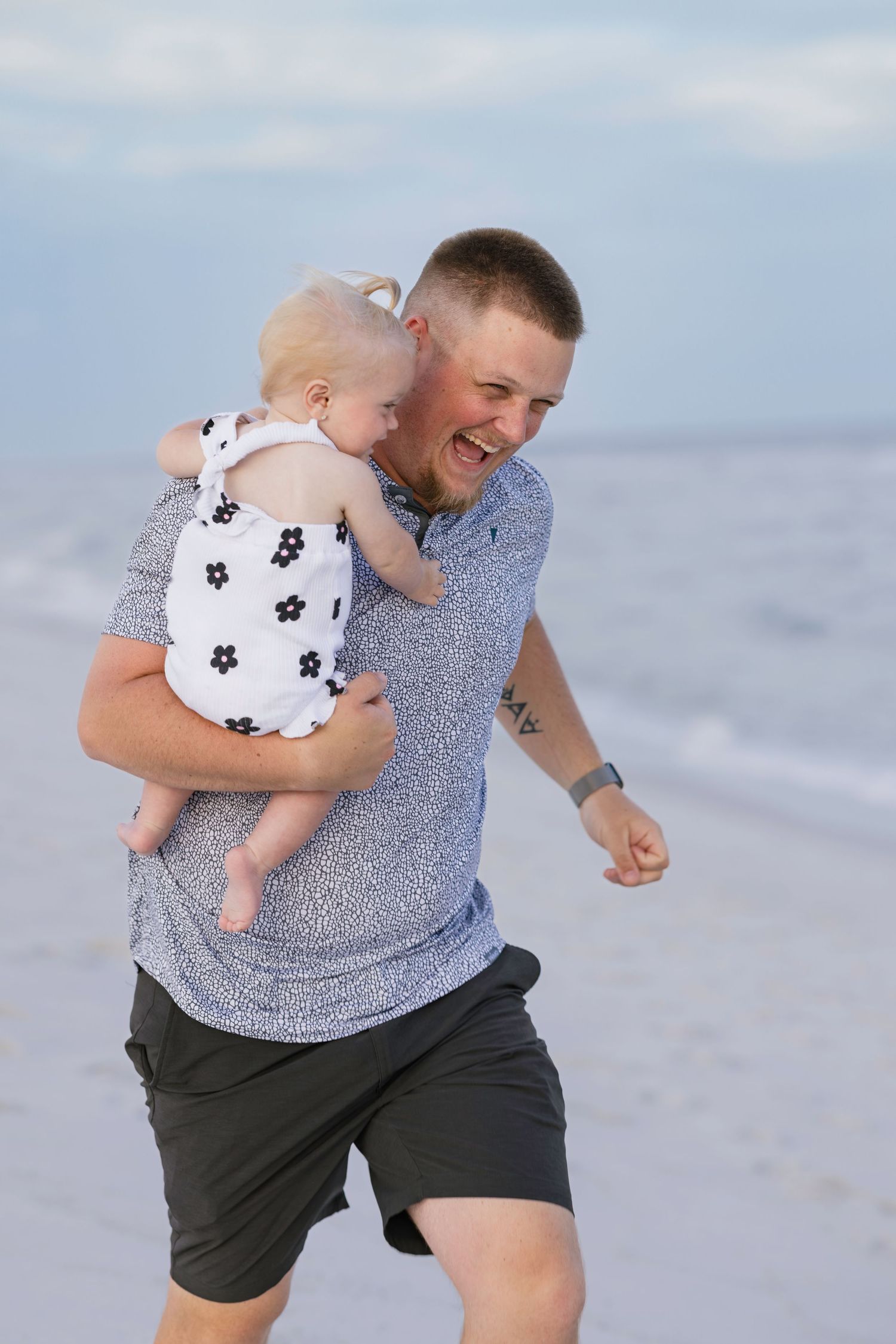Laughing father carrying baby on beach near ocean waves, wearing blue shirt and dark shorts.