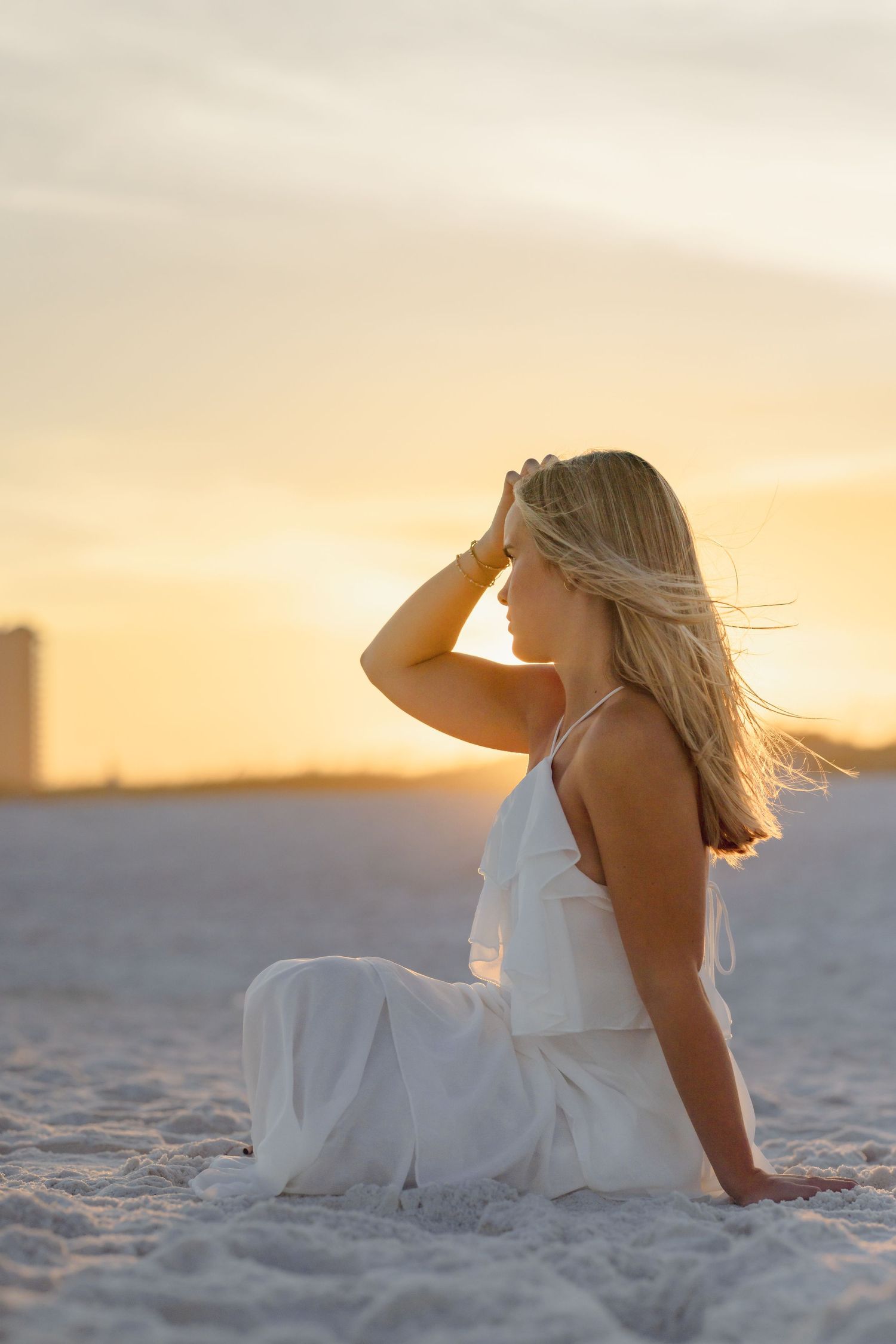 Blonde woman in white dress sitting on sandy beach at golden sunset, hand in hair, with serene ocean backdrop.