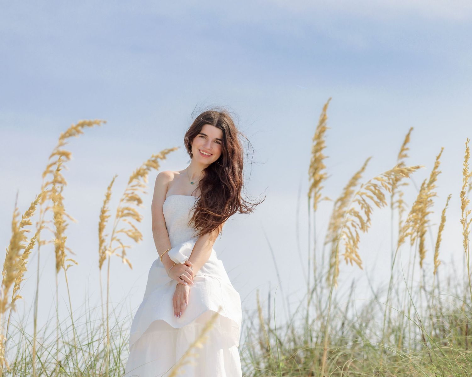 Young woman in white dress smiling among sea oats on a breezy beach day.