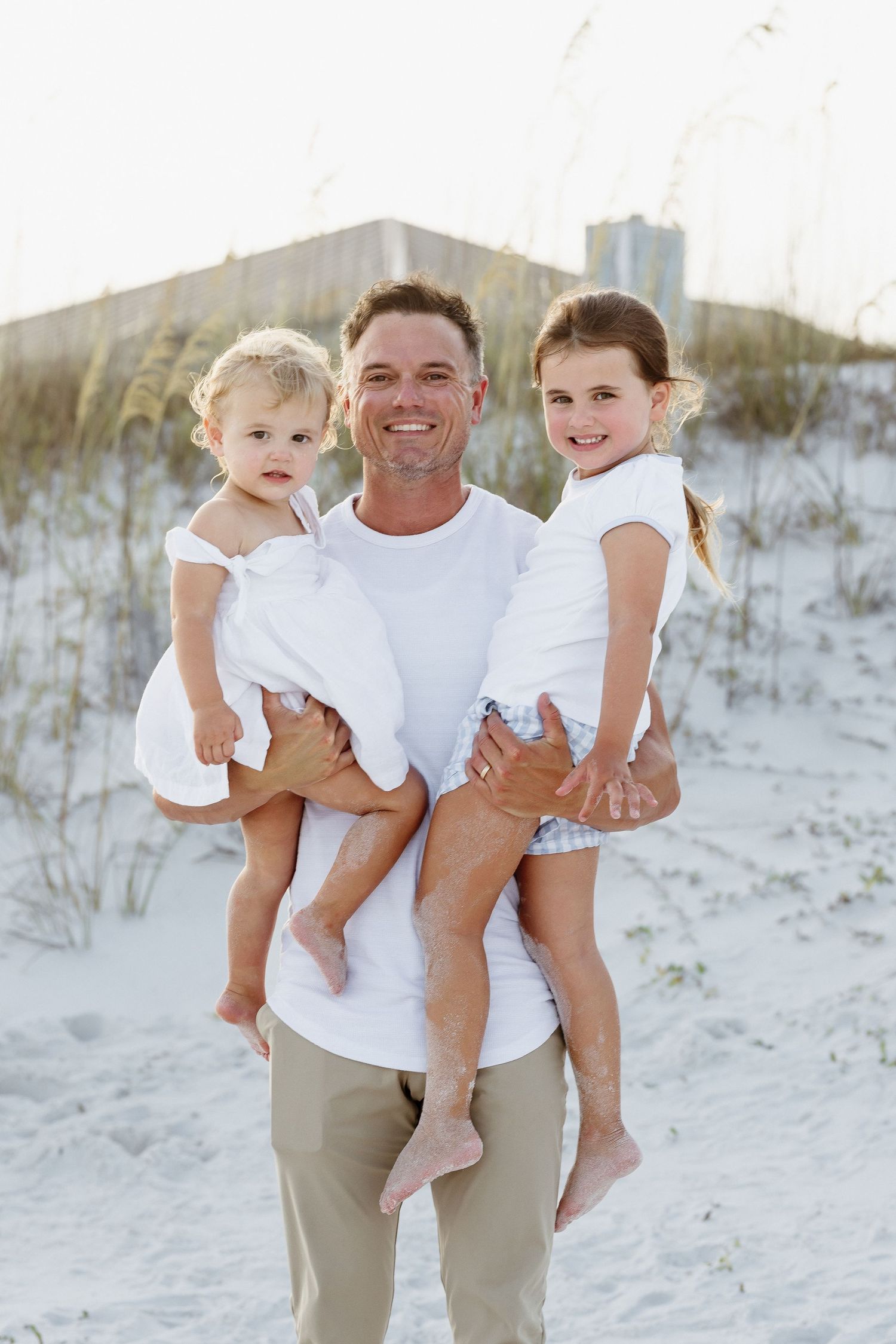 A smiling man holds two young girls in white dresses on a sandy beach with dunes in the background.