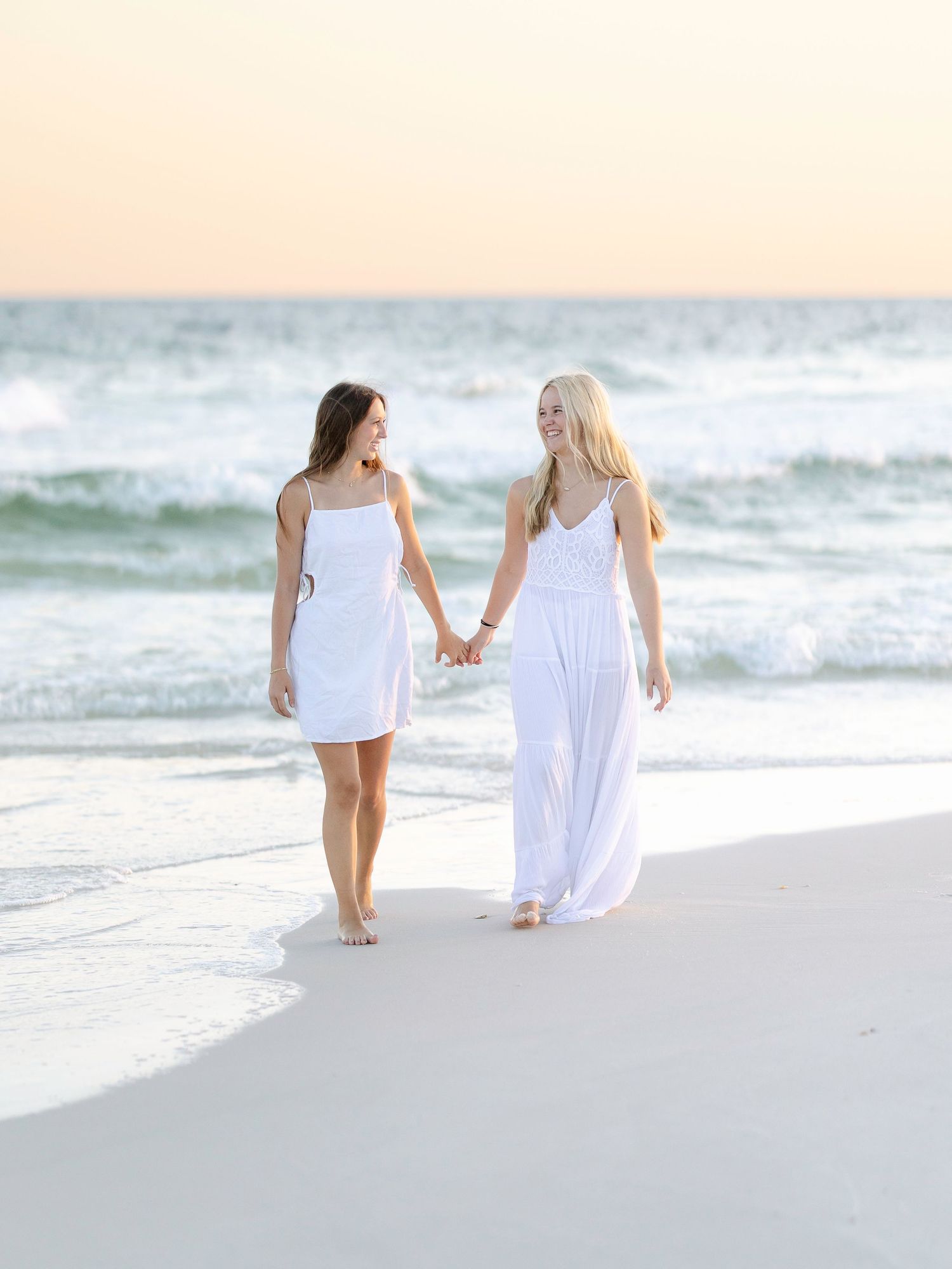Friends in flowing white dresses strolling together on beach near ocean waves.