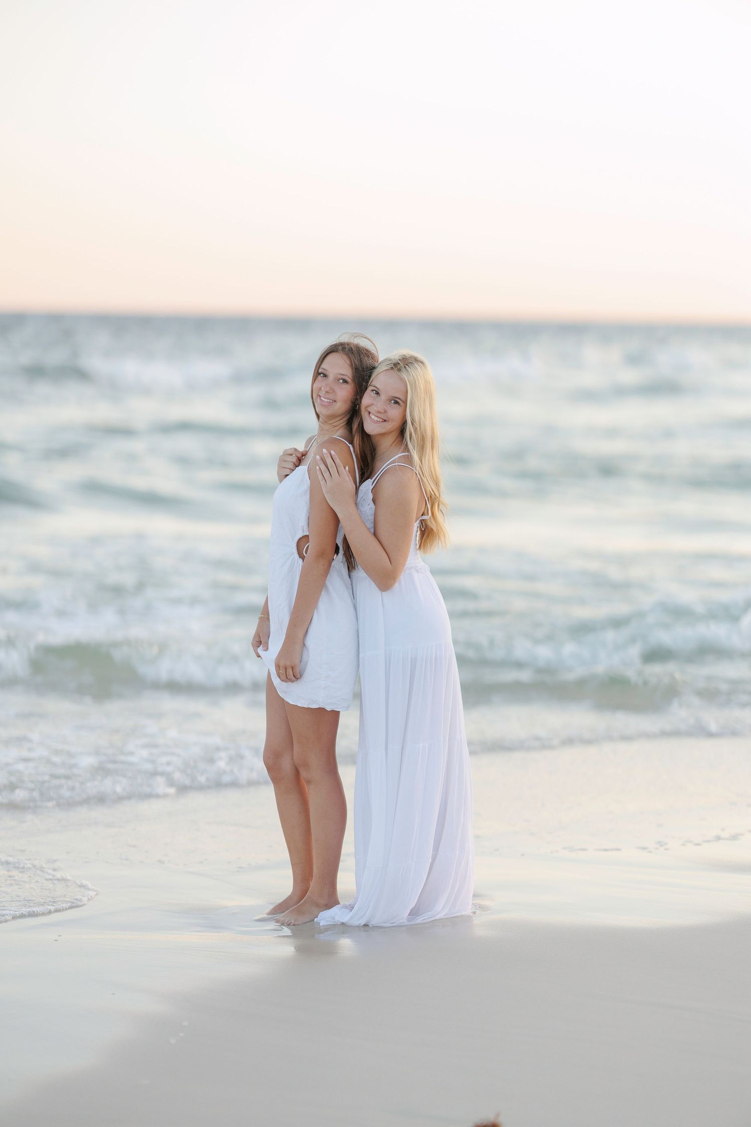 Two friends in white dresses embrace while standing on beach at sunset.