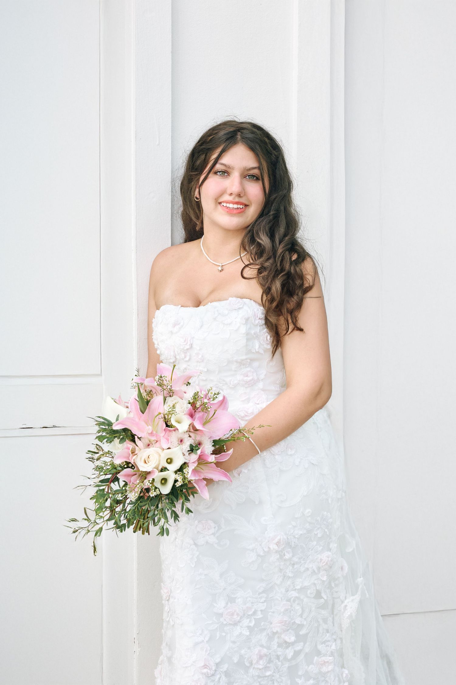 Bride in lace strapless wedding gown holds pink and white floral bouquet against white wall.