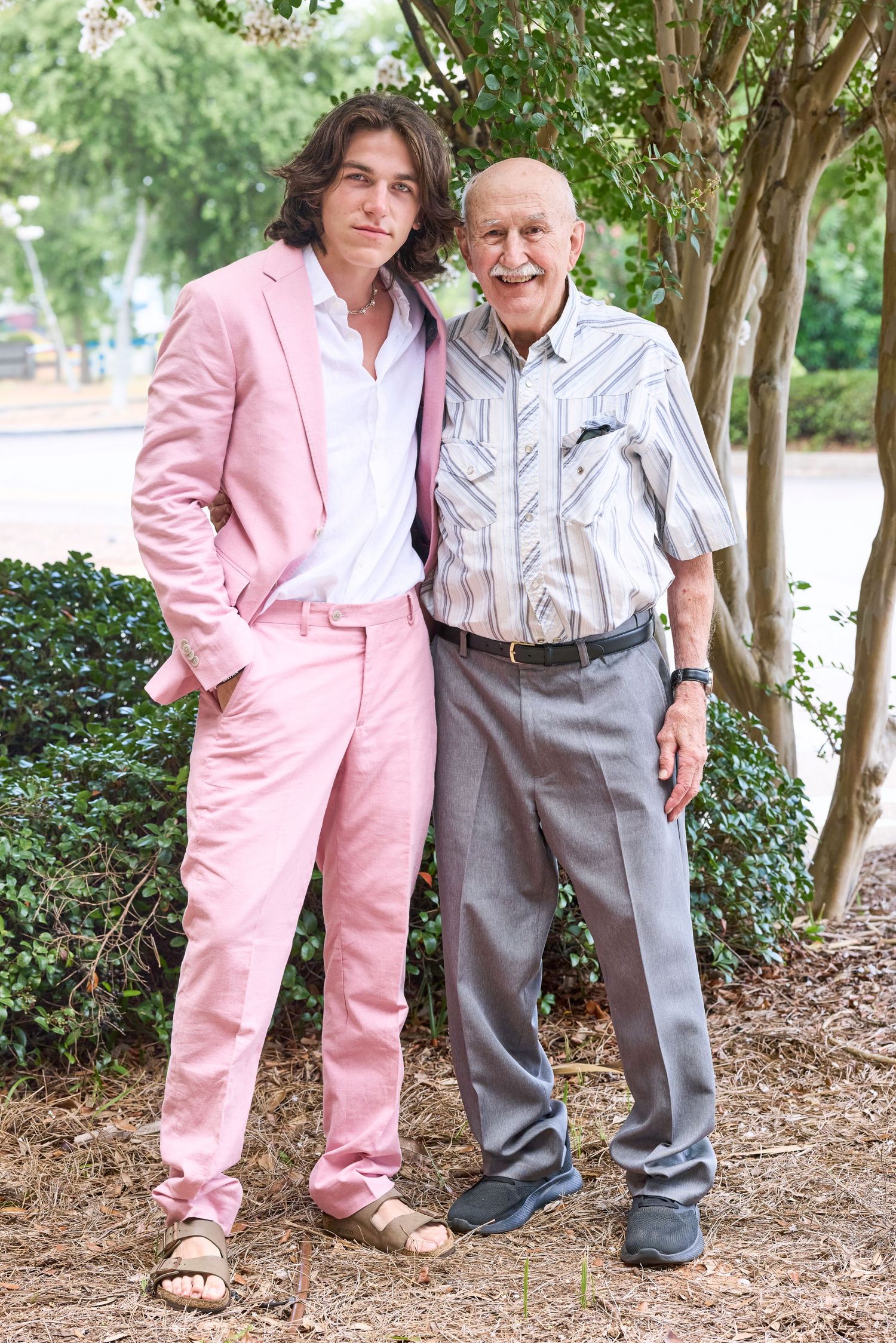 A pair in contrasting outfits pose together outdoors, one in a pink suit and one in casual gray attire.