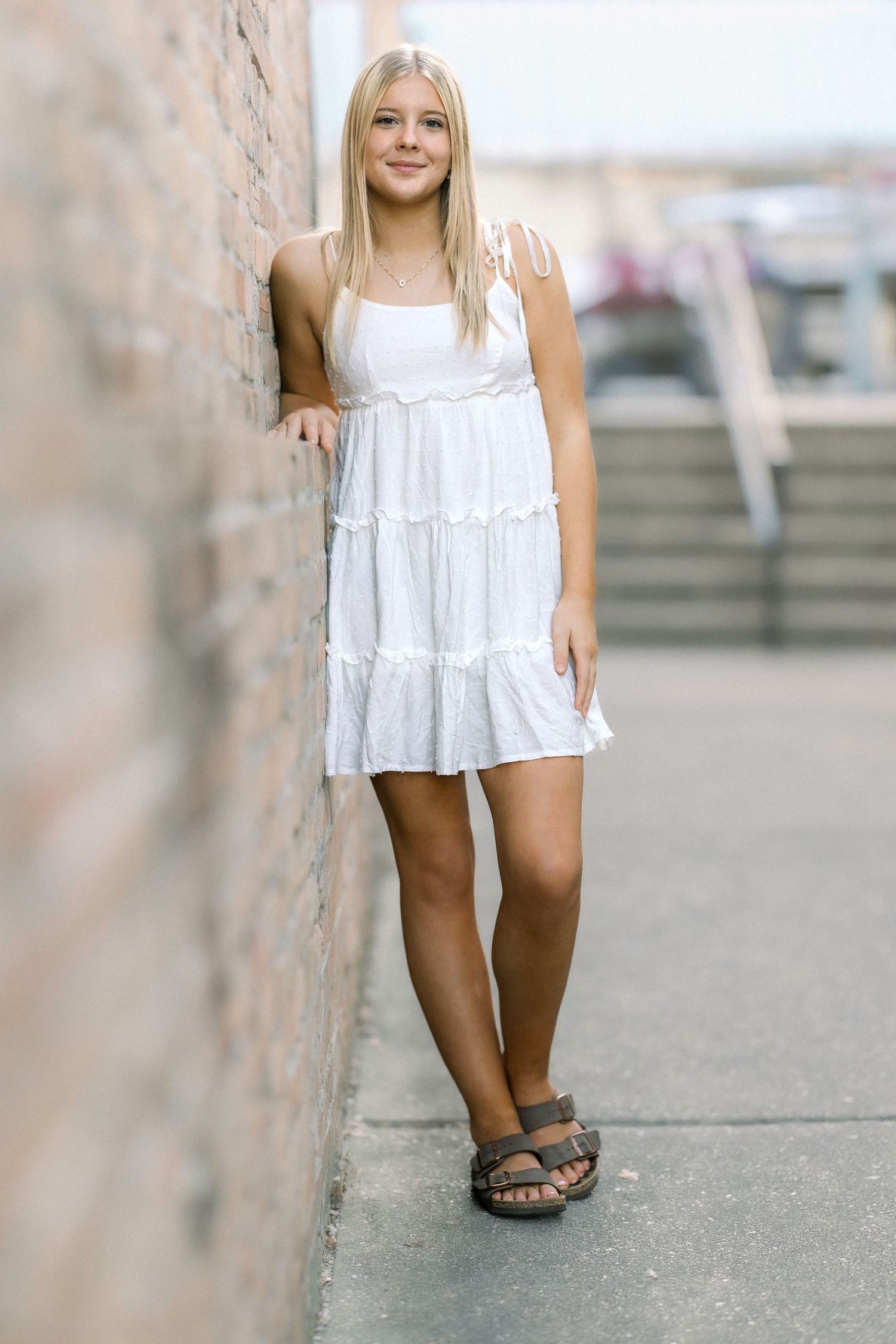Young blonde woman in white tiered mini dress leaning against brick wall outdoors.