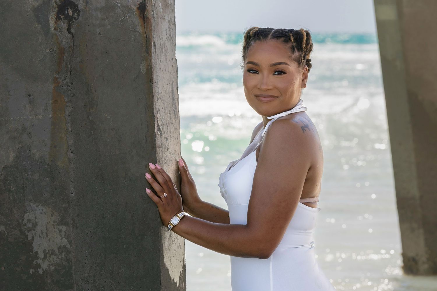 Young woman in white outfit leaning against pier pillar at sunny beach.