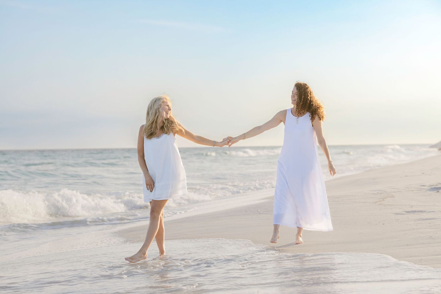 Two women in white dresses holding hands on a sunny beach with ocean waves in the background.