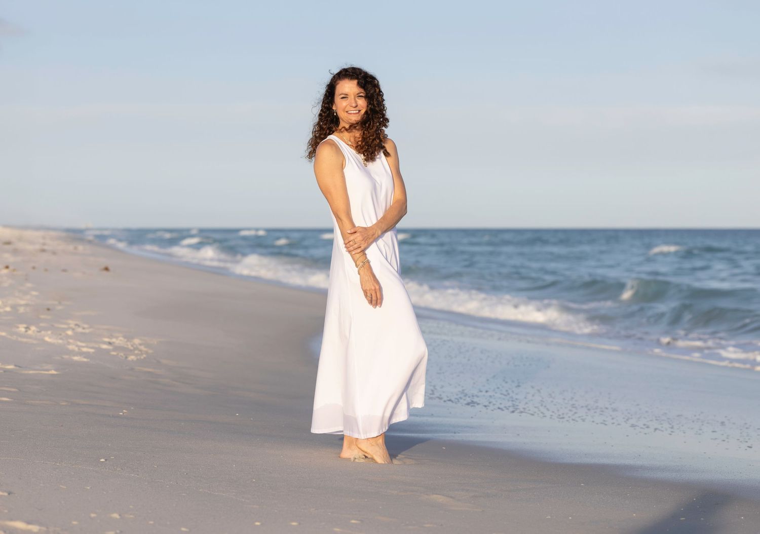 Woman in white maxi dress standing barefoot on sandy beach with ocean waves in background on clear day.
