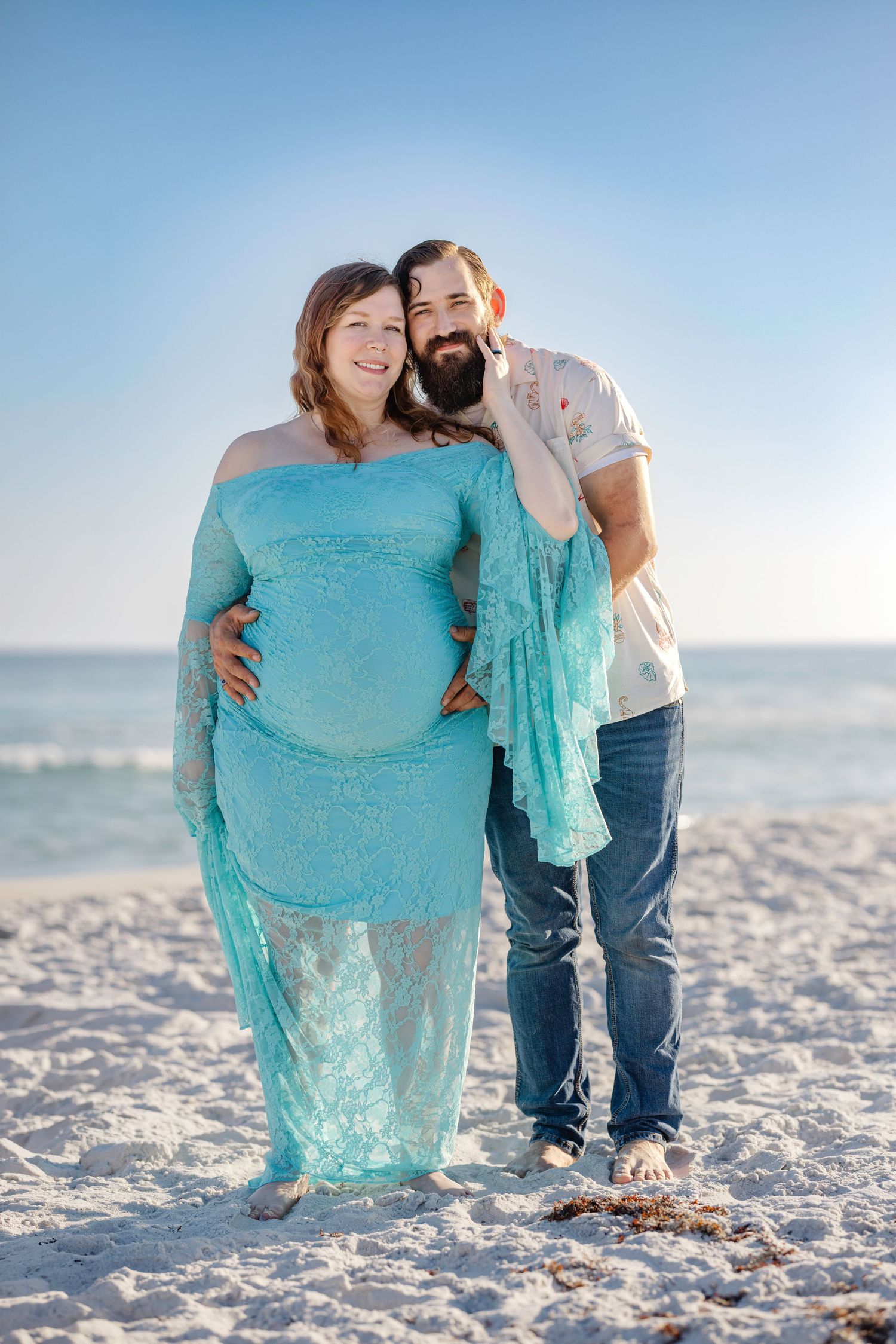 Pregnant woman in teal lace gown with bearded man on sunny beach, maternity photo shoot.