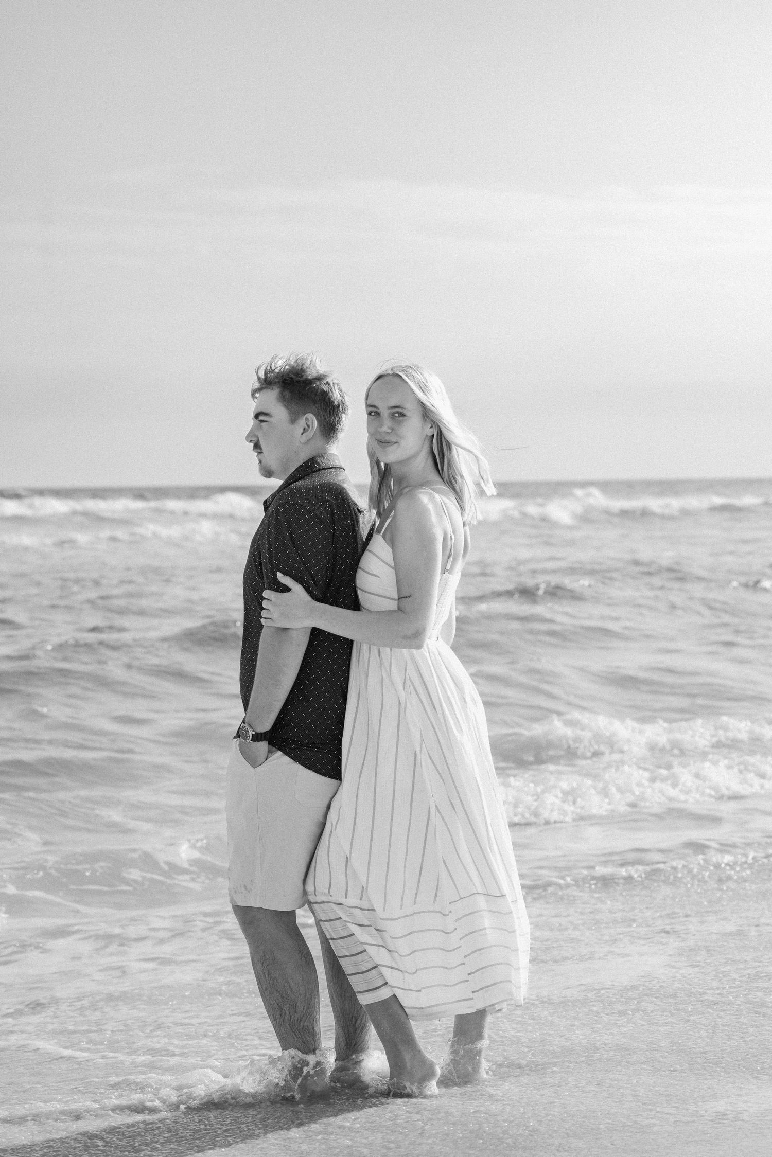 Black and white photo of a couple embracing on a beach with ocean waves behind them.