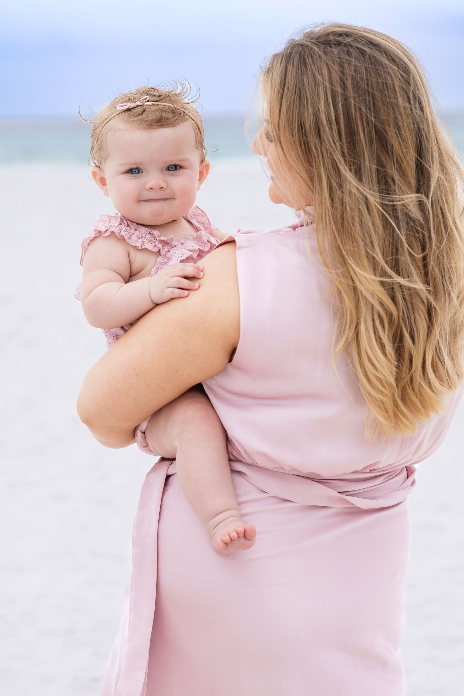 A mother in a pink dress holds her baby girl on a sunny beach, with the infant gazing forward sweetly.