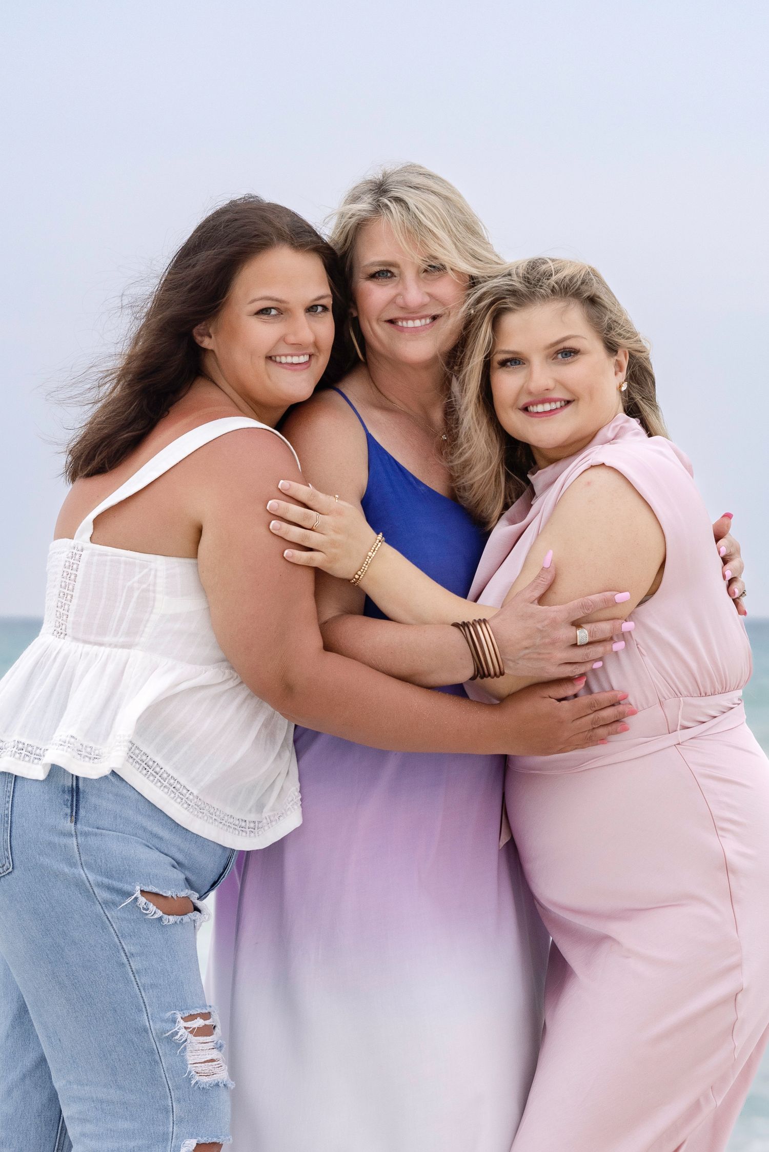 Three smiling women in colorful summer outfits hugging together on a beach with ocean in the background.