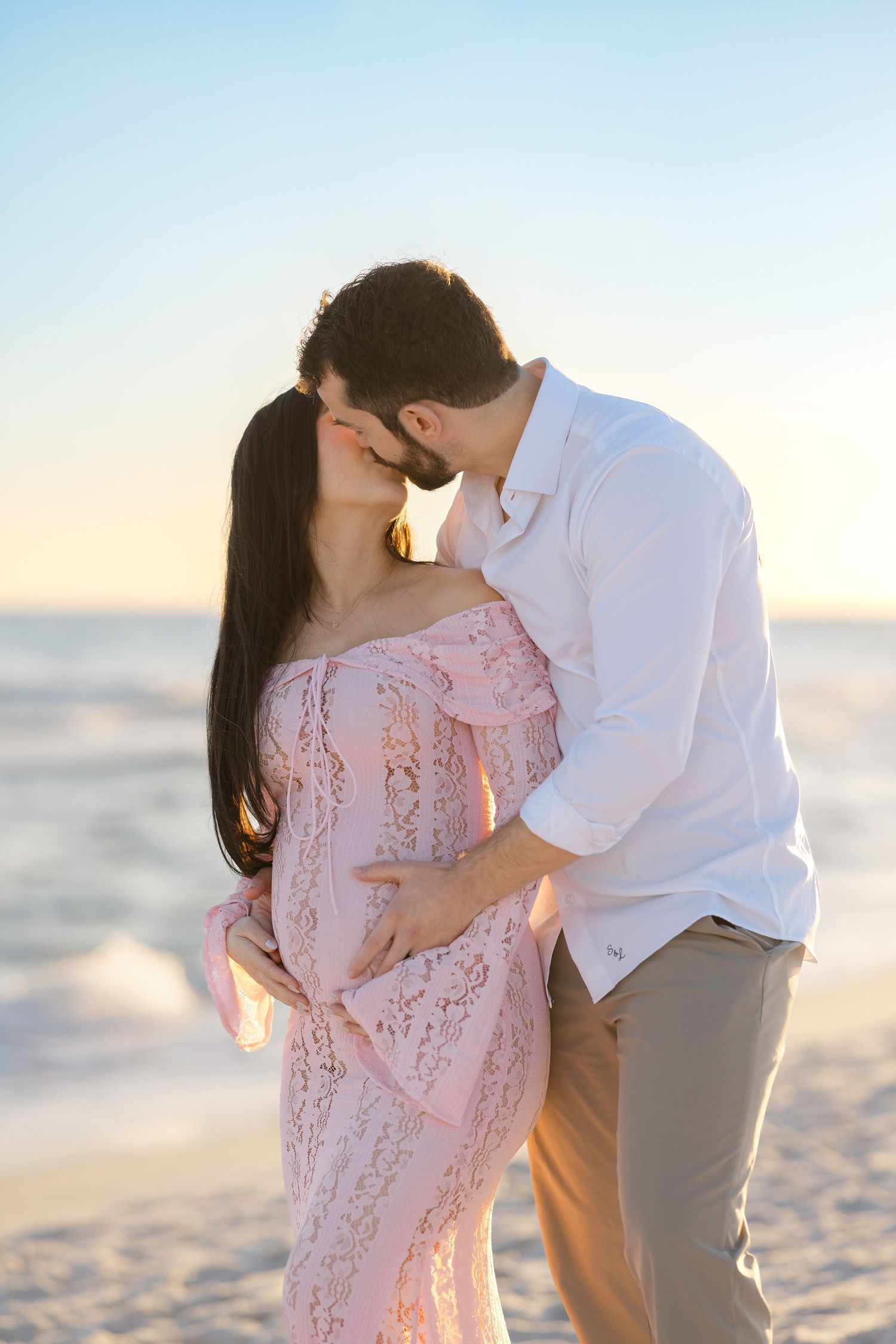 Romantic couple embraces on a sandy beach at sunset wearing pink and white outfits.