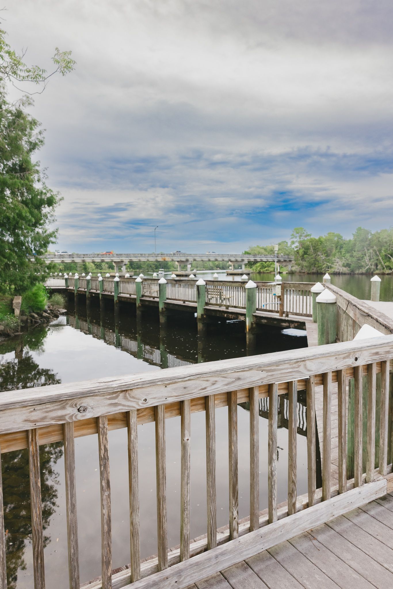 Wooden boardwalk dock extending over calm water with lush green trees and cloudy sky reflected in the serene waterway.