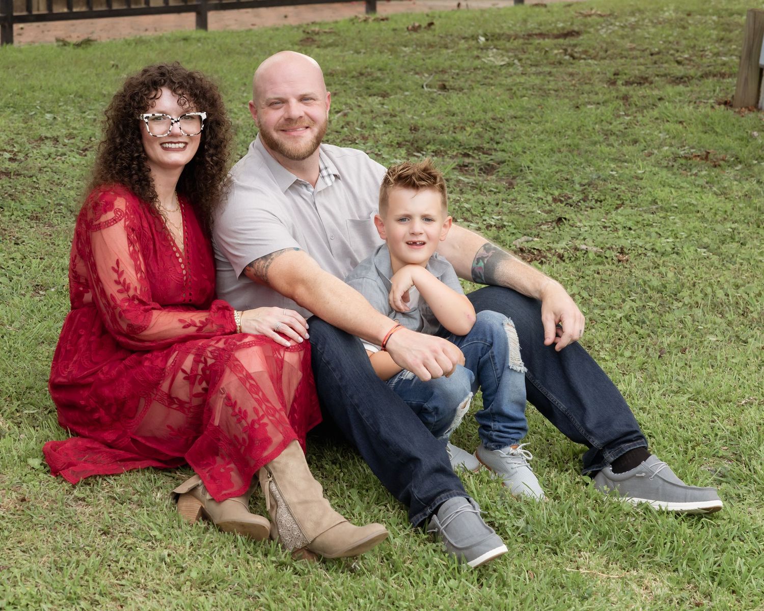 A smiling family of three sits on green grass outdoors, woman in red dress, man in gray shirt, and young boy in jeans.
