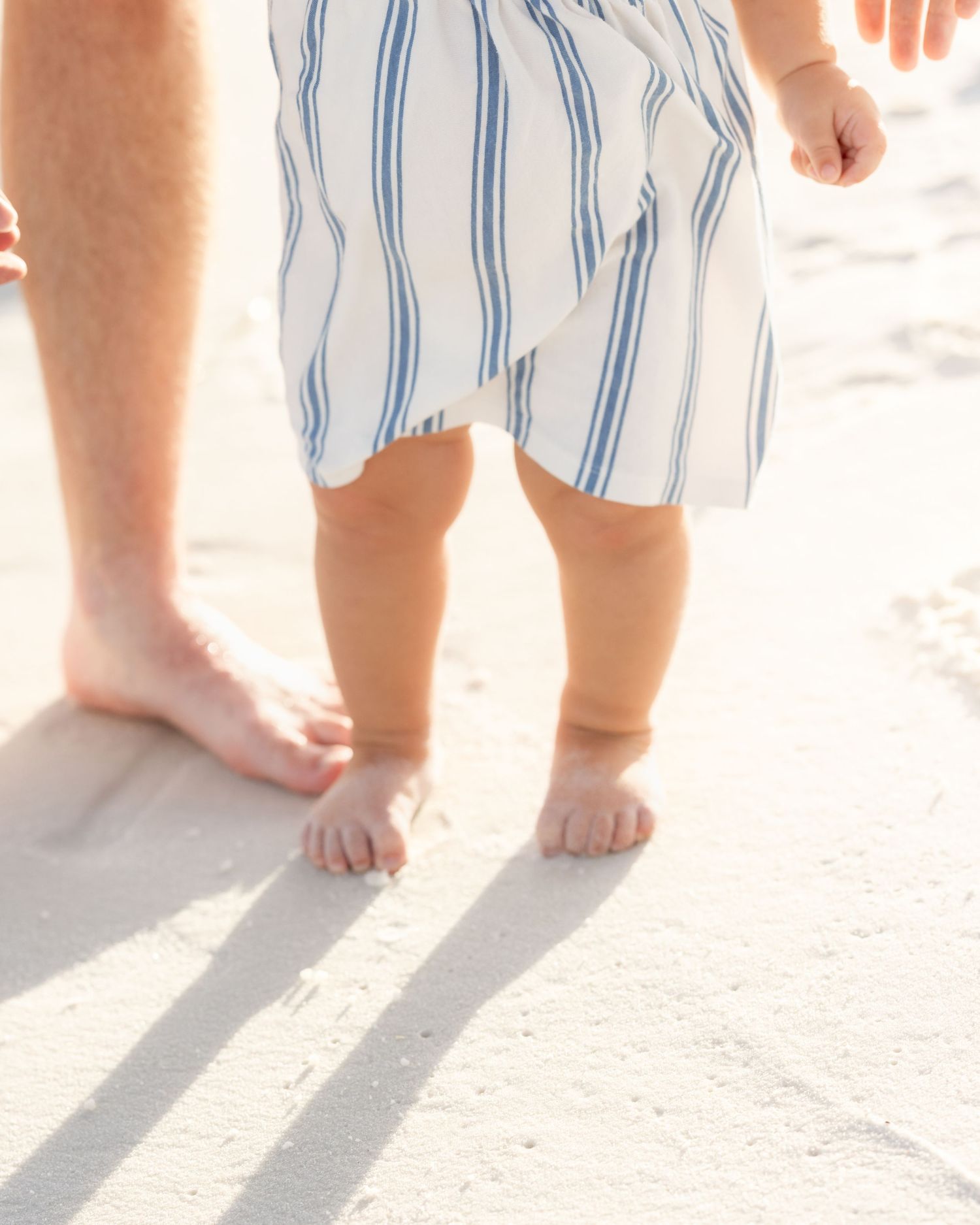 A toddler in a striped dress stands barefoot on white sand beach beside an adult's bare feet.