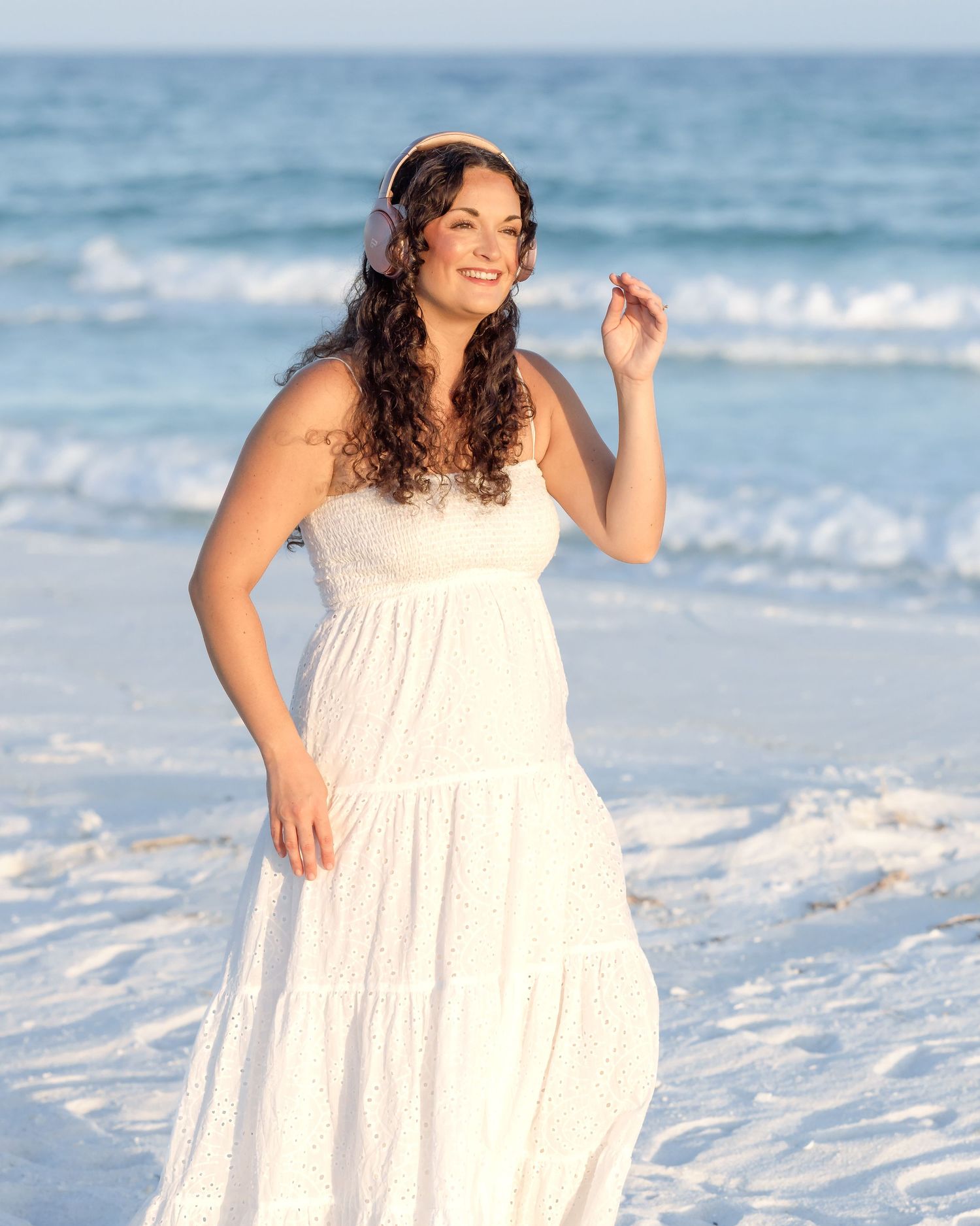 Woman in white tiered maxi dress wearing headphones smiles on a sunny beach with ocean waves behind her.