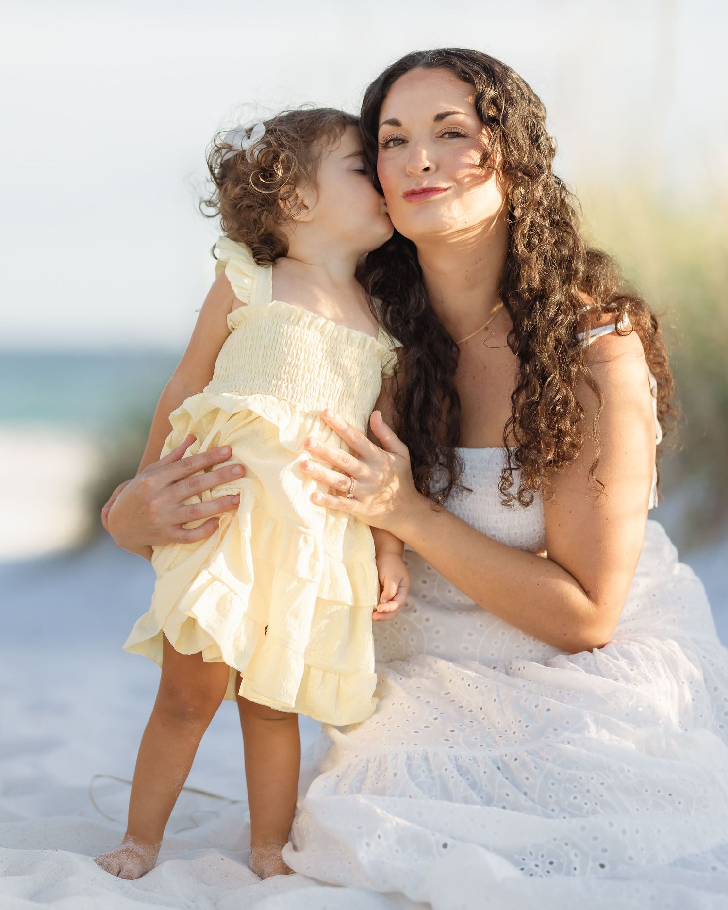A young girl in a yellow dress kisses her mother on the cheek while sitting on a sandy beach.