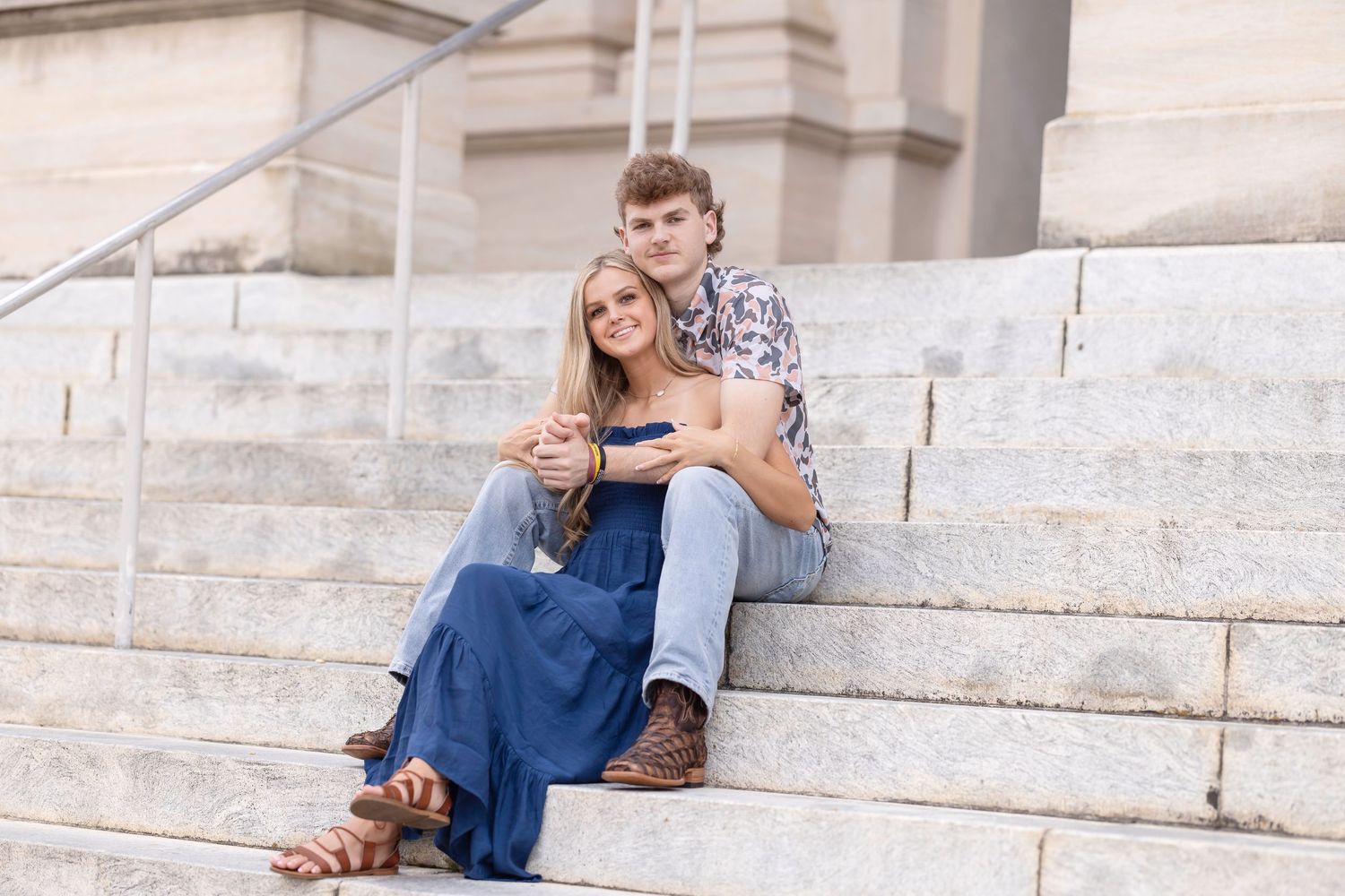 A couple sits together on white stone steps, with the woman wearing a blue dress and the man in casual attire.