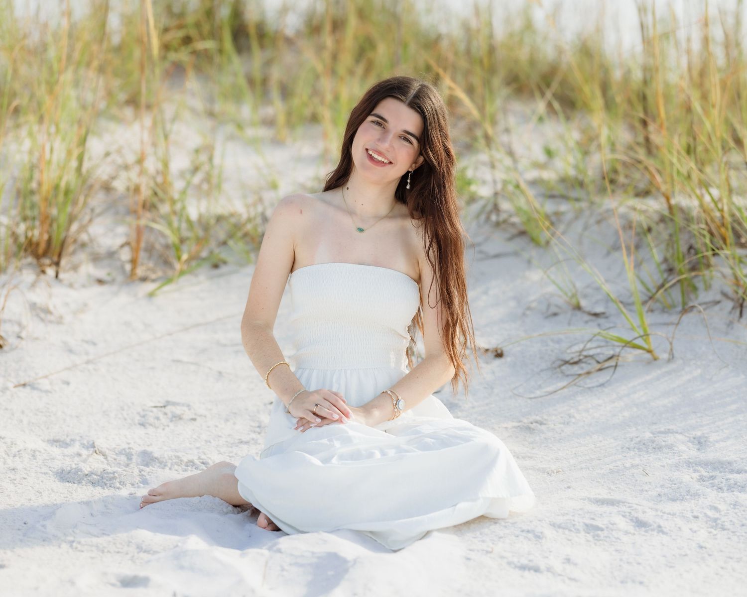 Young woman in white strapless dress sitting on sandy beach dunes, smiling with long brown hair, golden hour light.
