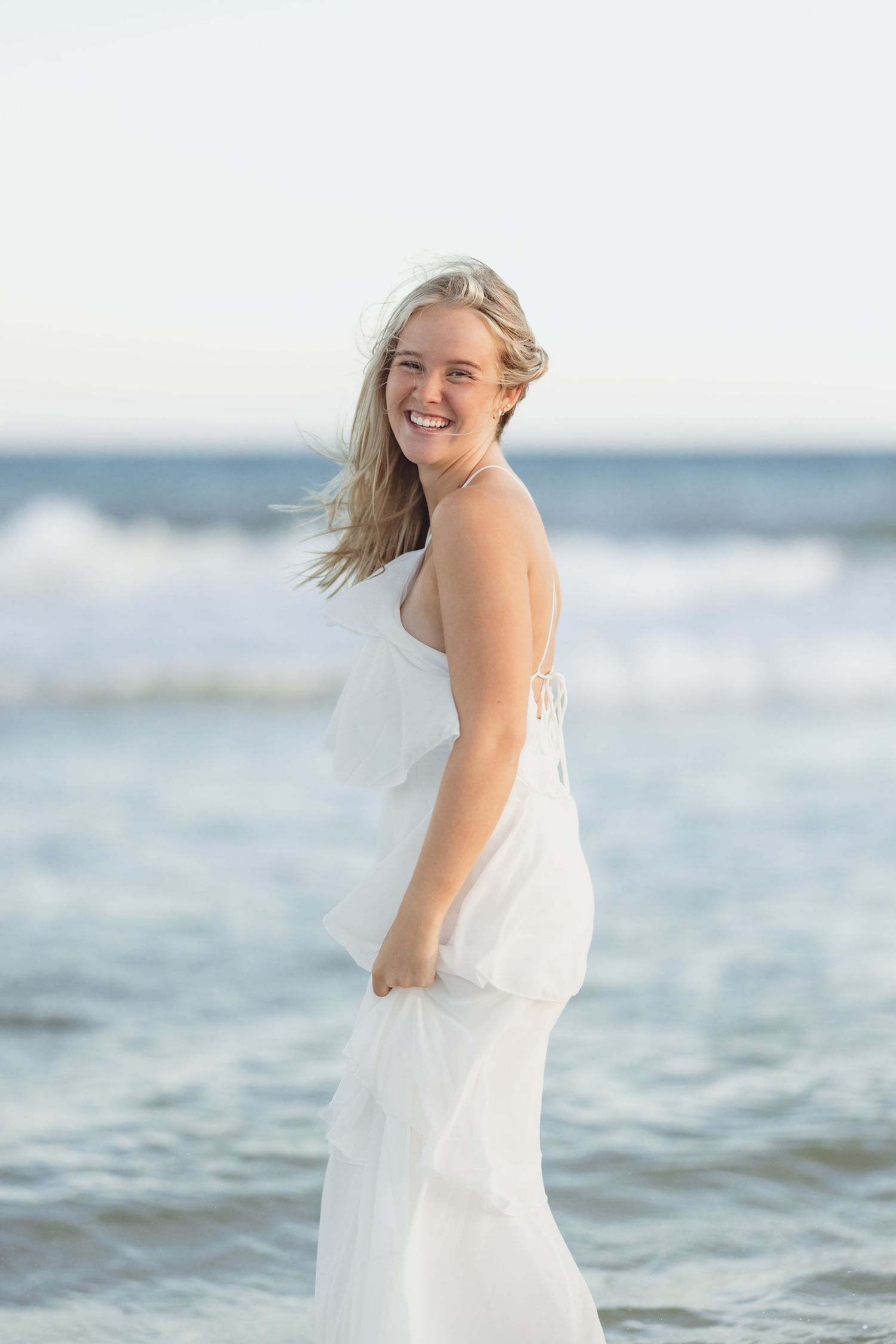Smiling blonde woman in white dress standing at ocean shoreline with waves.