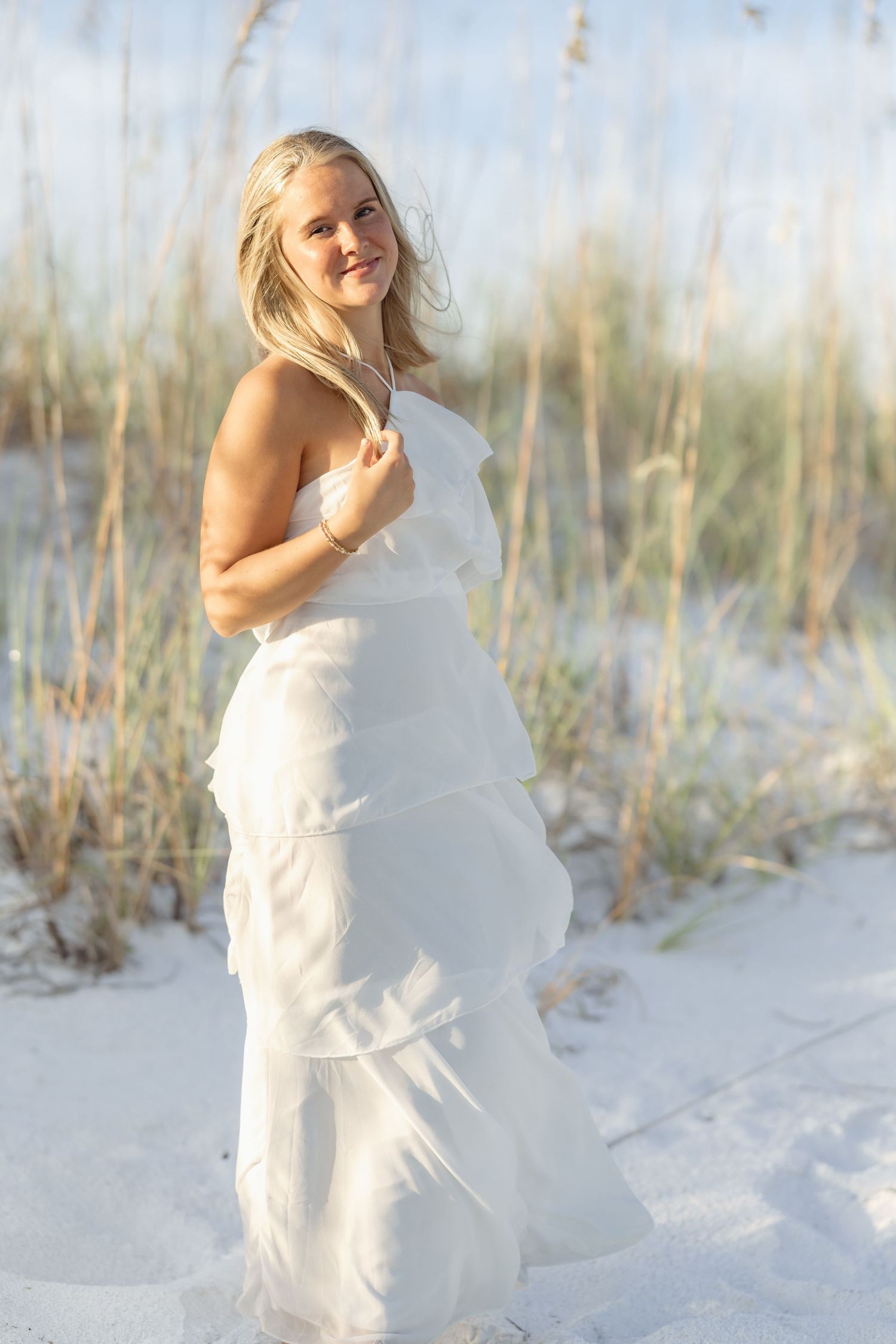 Blonde woman in white tiered maxi dress posing on white sand beach dunes with sea oats in background.