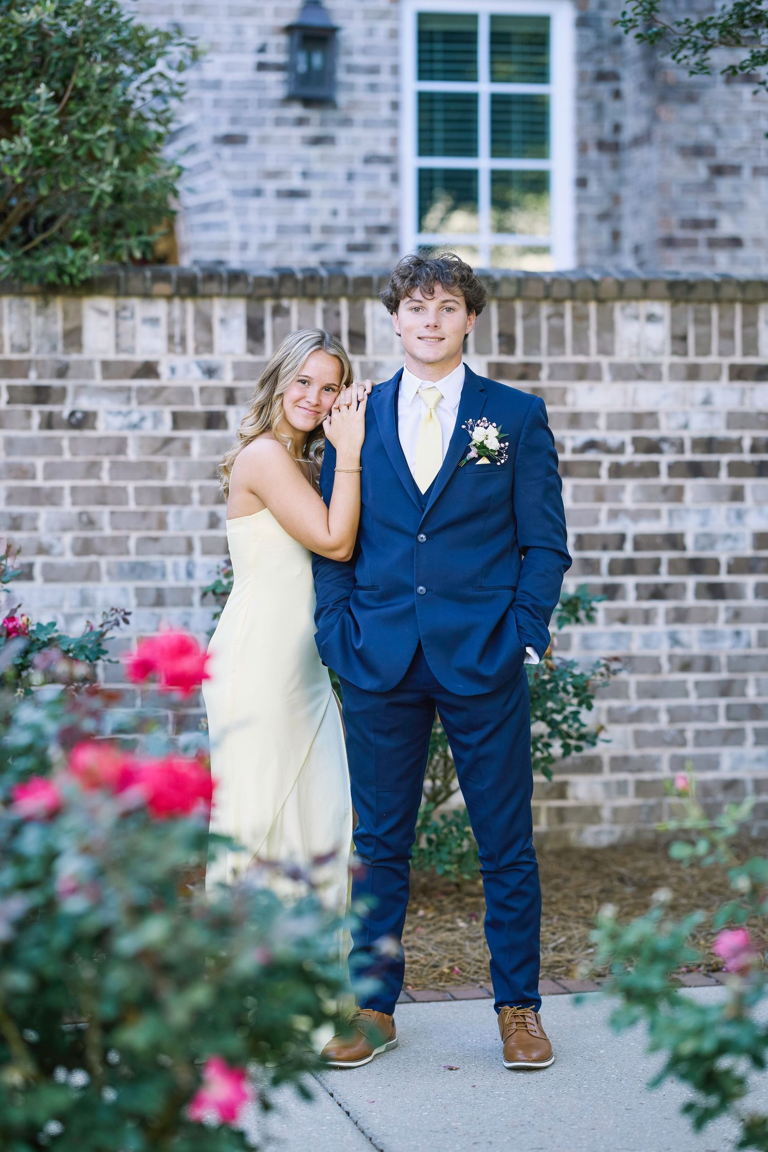 A couple in formal attire poses against a brick wall with blooming rose bushes in the foreground.